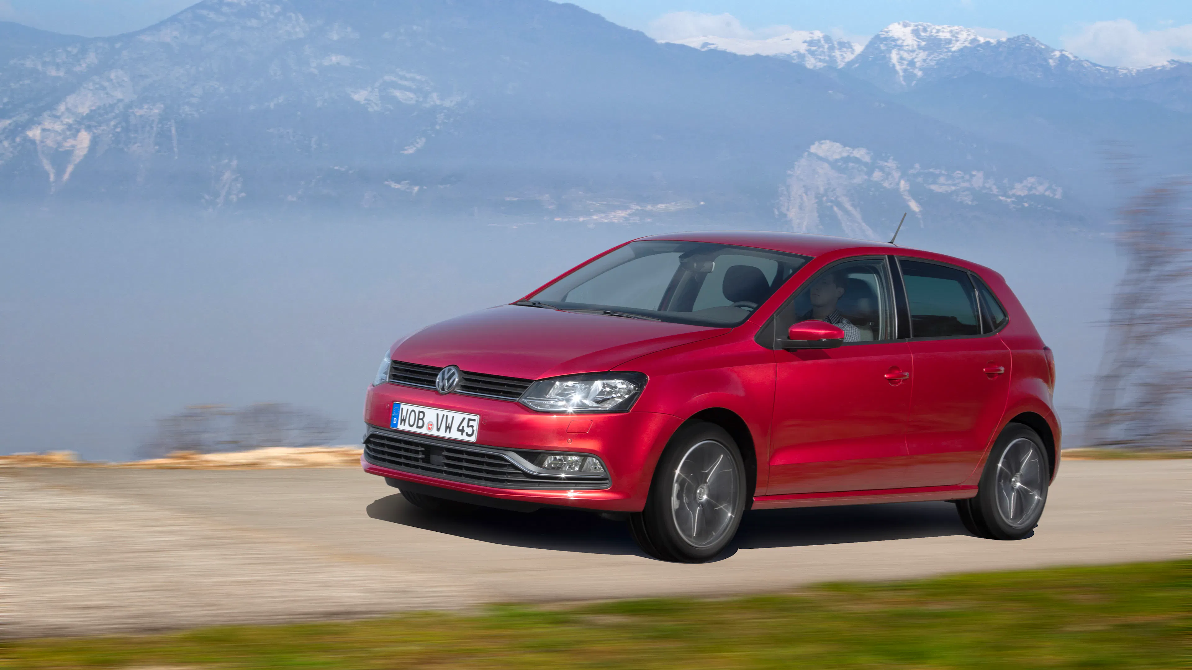Roter VW Polo V mit Facelift-Front fährt vor Bergpanorama mit Schneegipfeln über eine Schotterstraße.