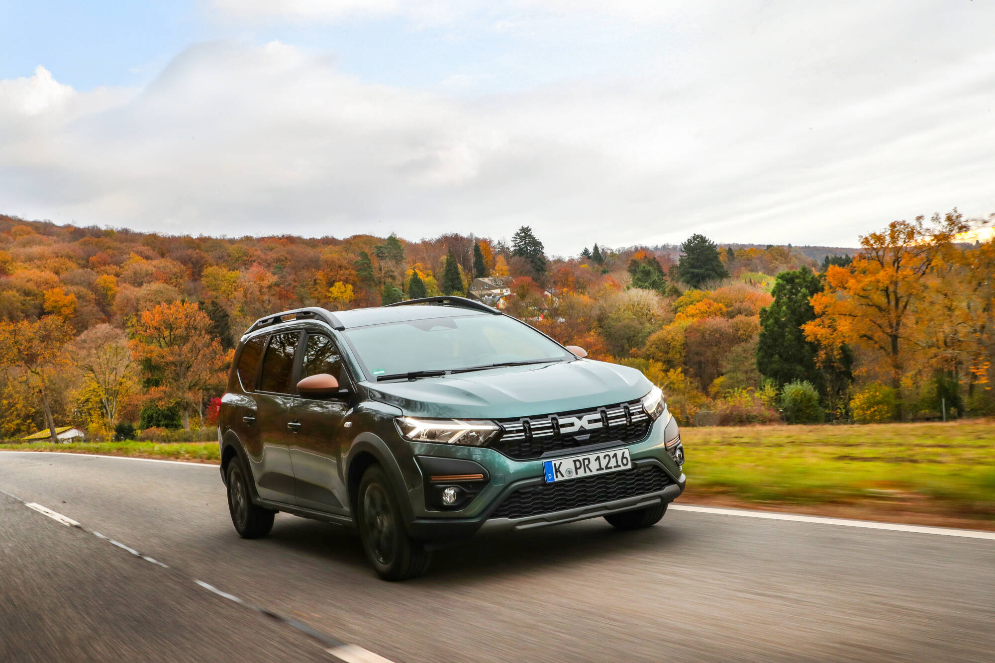 Grüner Dacia Jogger fährt frontal auf einer Landstraße vor einem herbstlich gefärbten Wald.