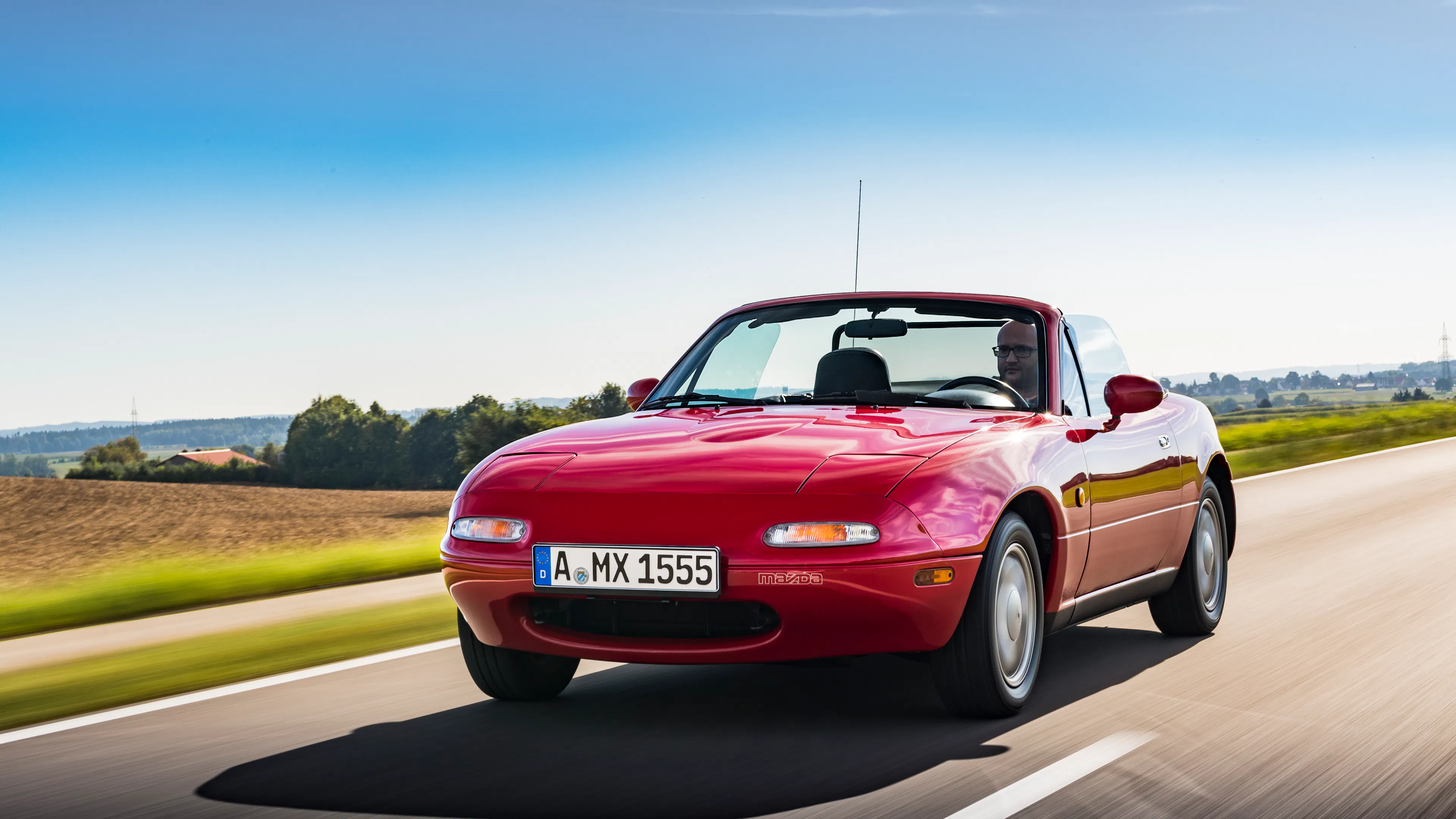 Roter Mazda MX-5 NA Roadster mit offenem Verdeck fährt auf einer Landstraße, Frontansicht mit Bewegungsunschärfe im Hintergrund.