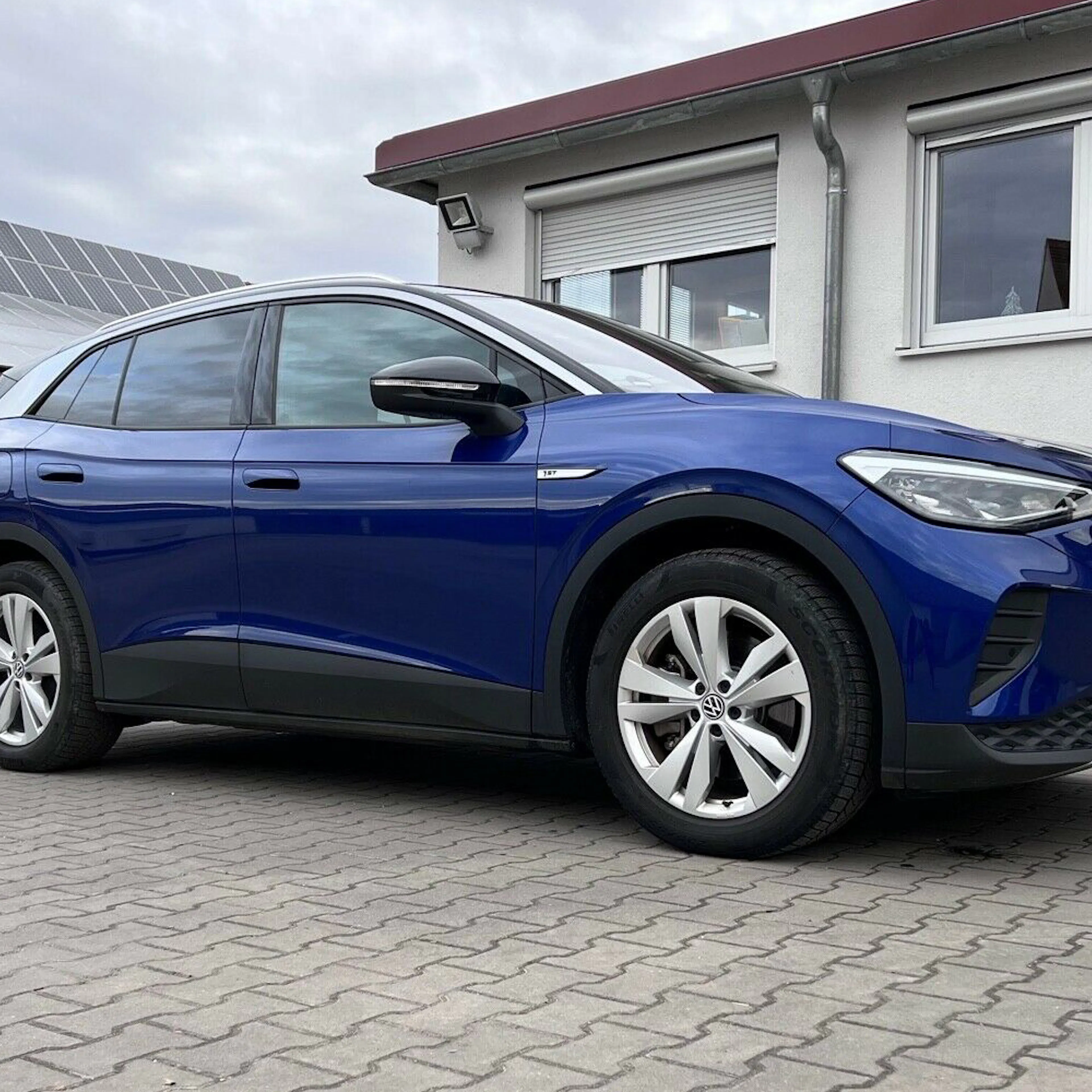 Blauer SUV, geparkt auf einem gepflasterten Hof vor einem Gebäude mit weißen Fenstern und Solarmodulen auf dem Dach.