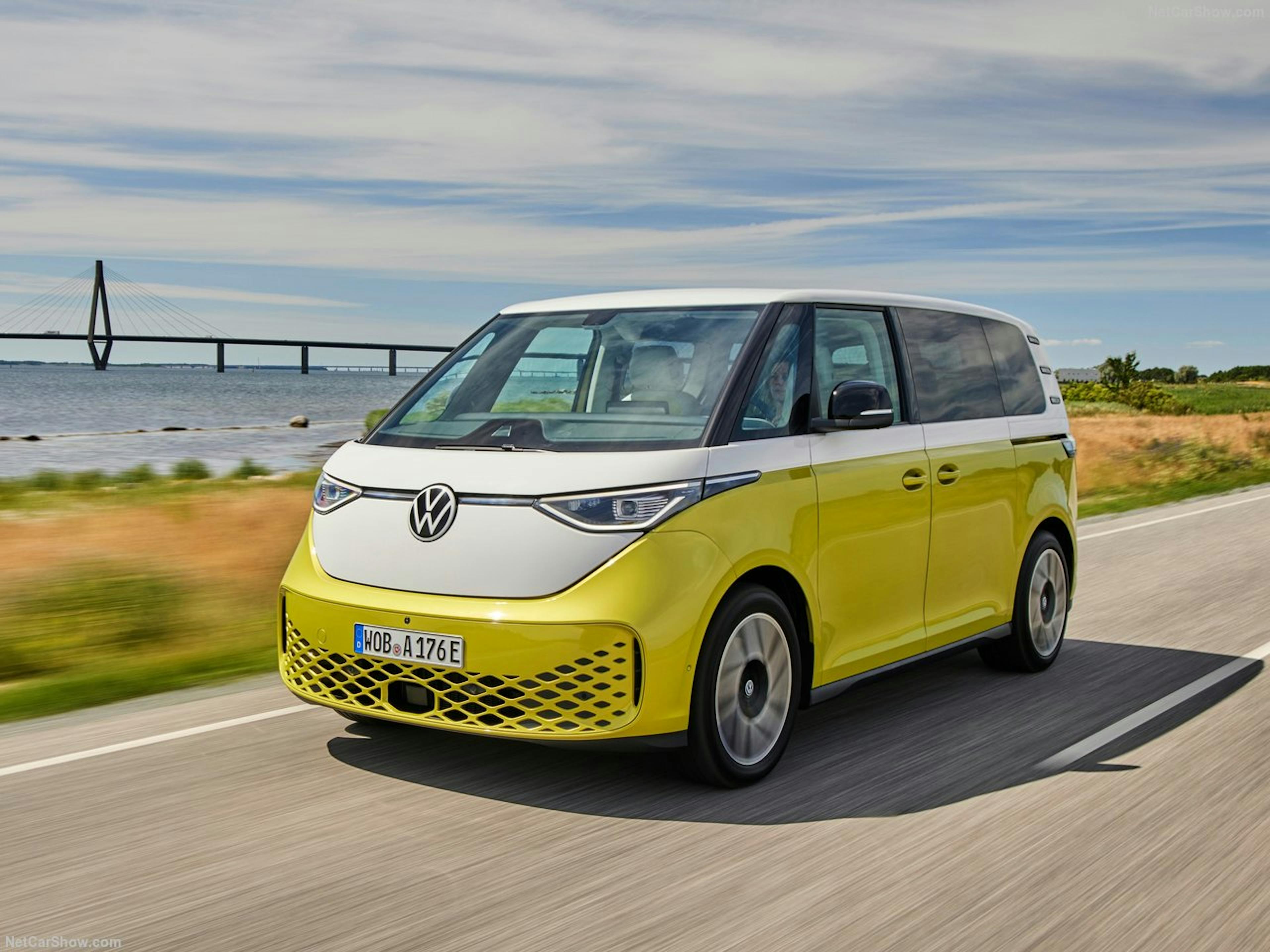 Ein gelb-weißer VW ID. Buzz fährt auf einer Landstraße am Meer entlang, im Hintergrund eine Brücke und blauer Himmel.