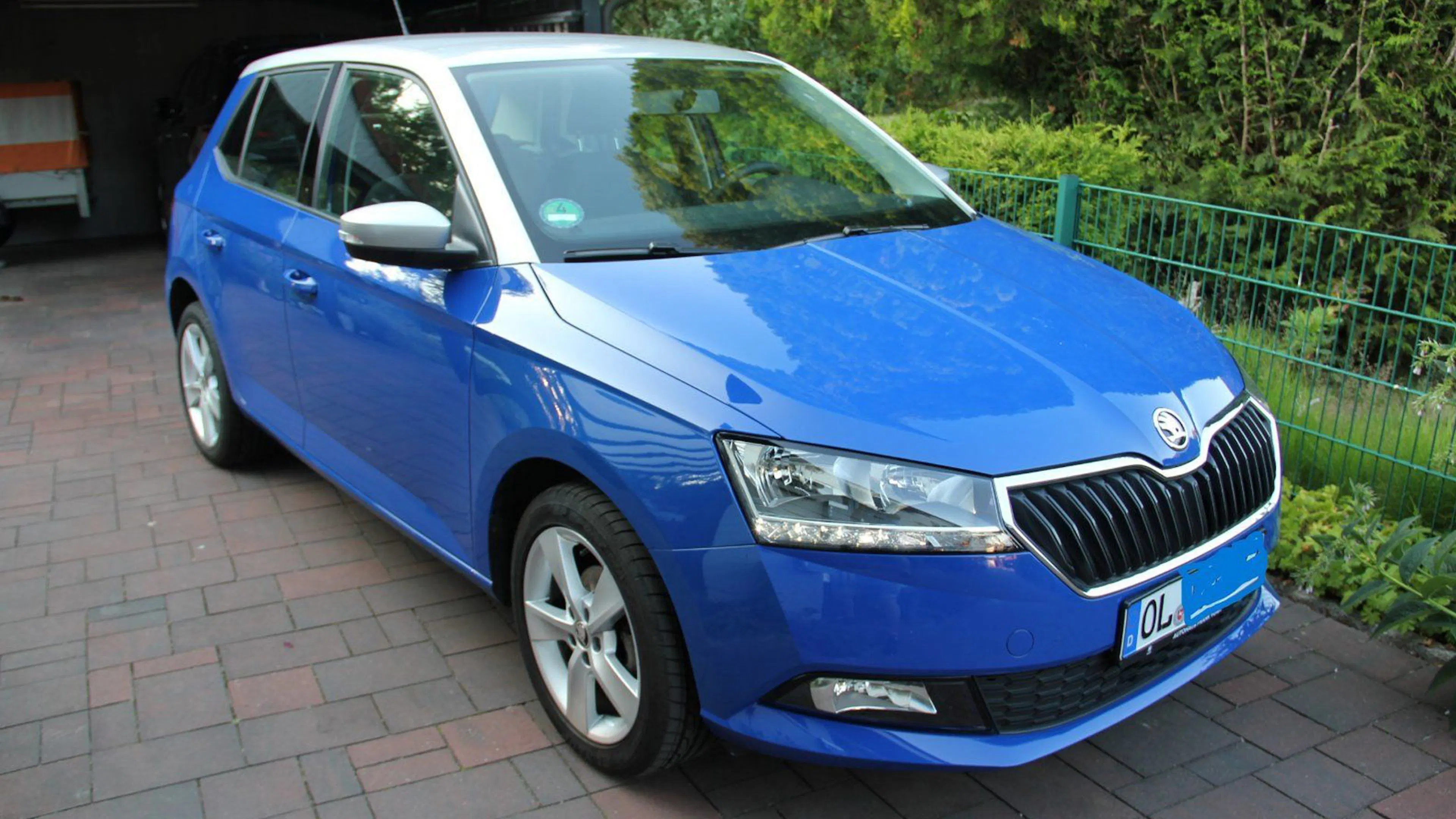 Blauer Skoda Fabia steht auf einer gepflasterten Einfahrt, umgeben von grüner Vegetation und einem Metallzaun im Hintergrund.