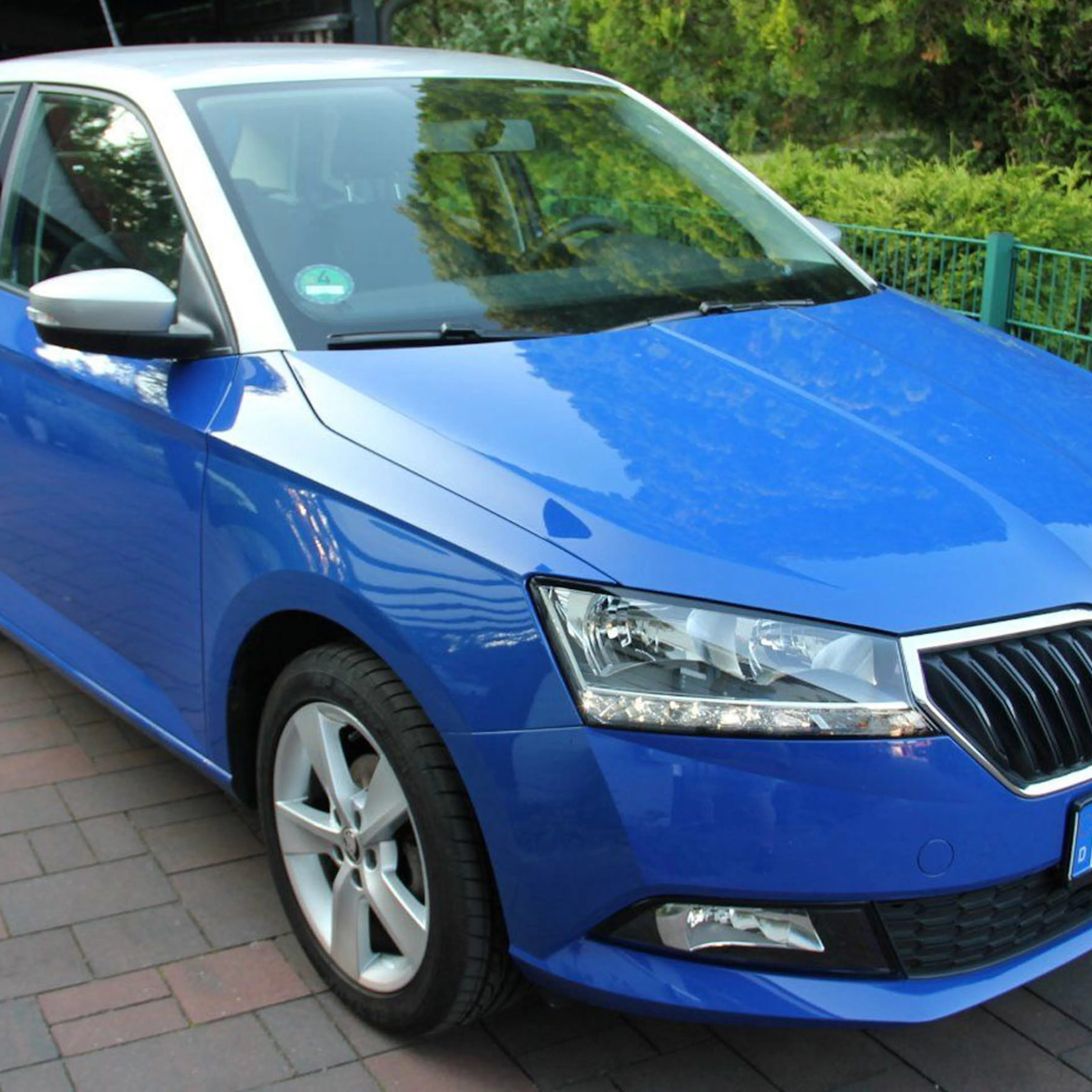 Blauer Skoda Fabia steht auf einer gepflasterten Einfahrt, umgeben von grüner Vegetation und einem Metallzaun im Hintergrund.