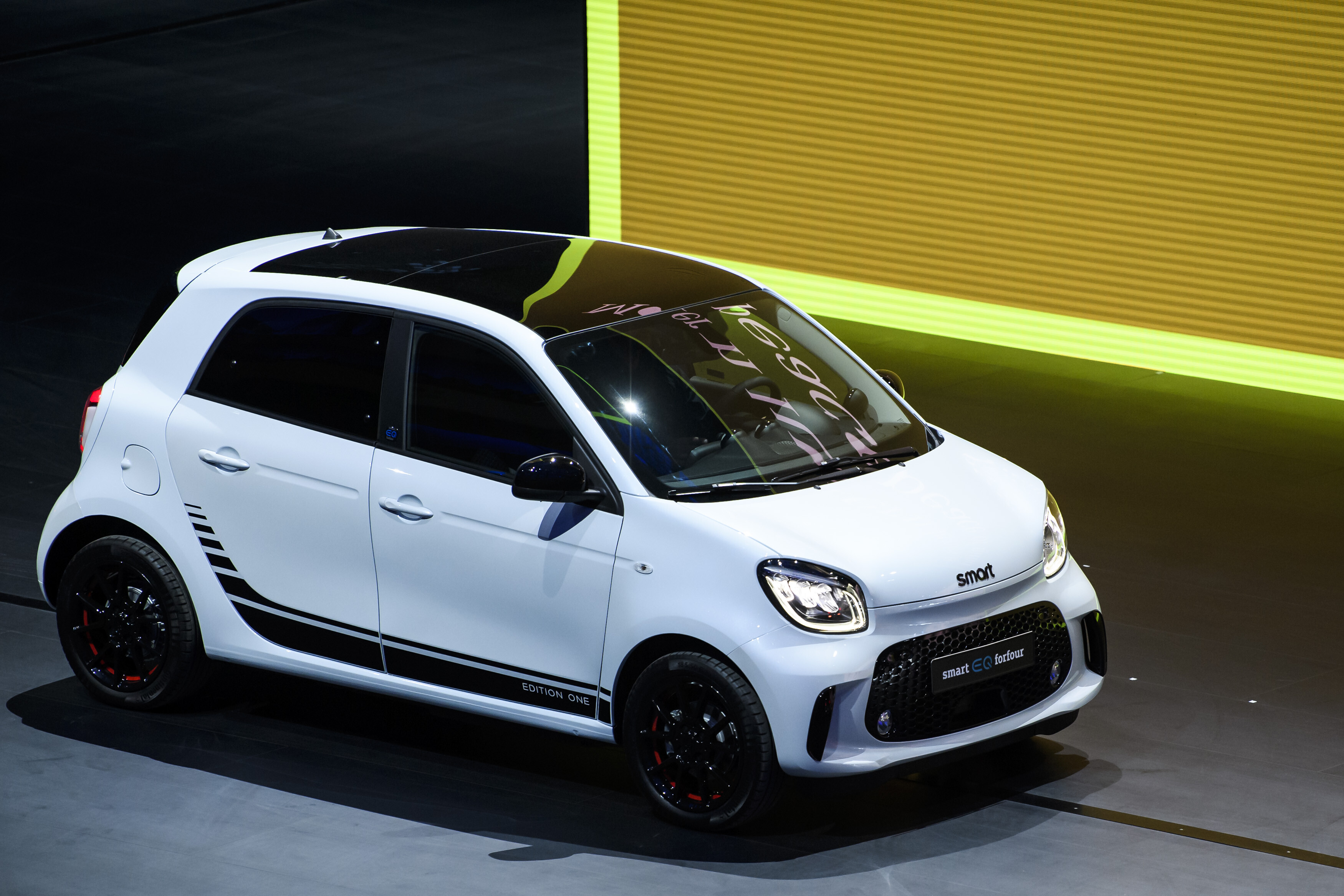 Weißer Smart EQ ForFour Edition One mit schwarzem Dach und schwarzen Felgen auf einer Bühne vor gelb-grünem Leuchtschirm.