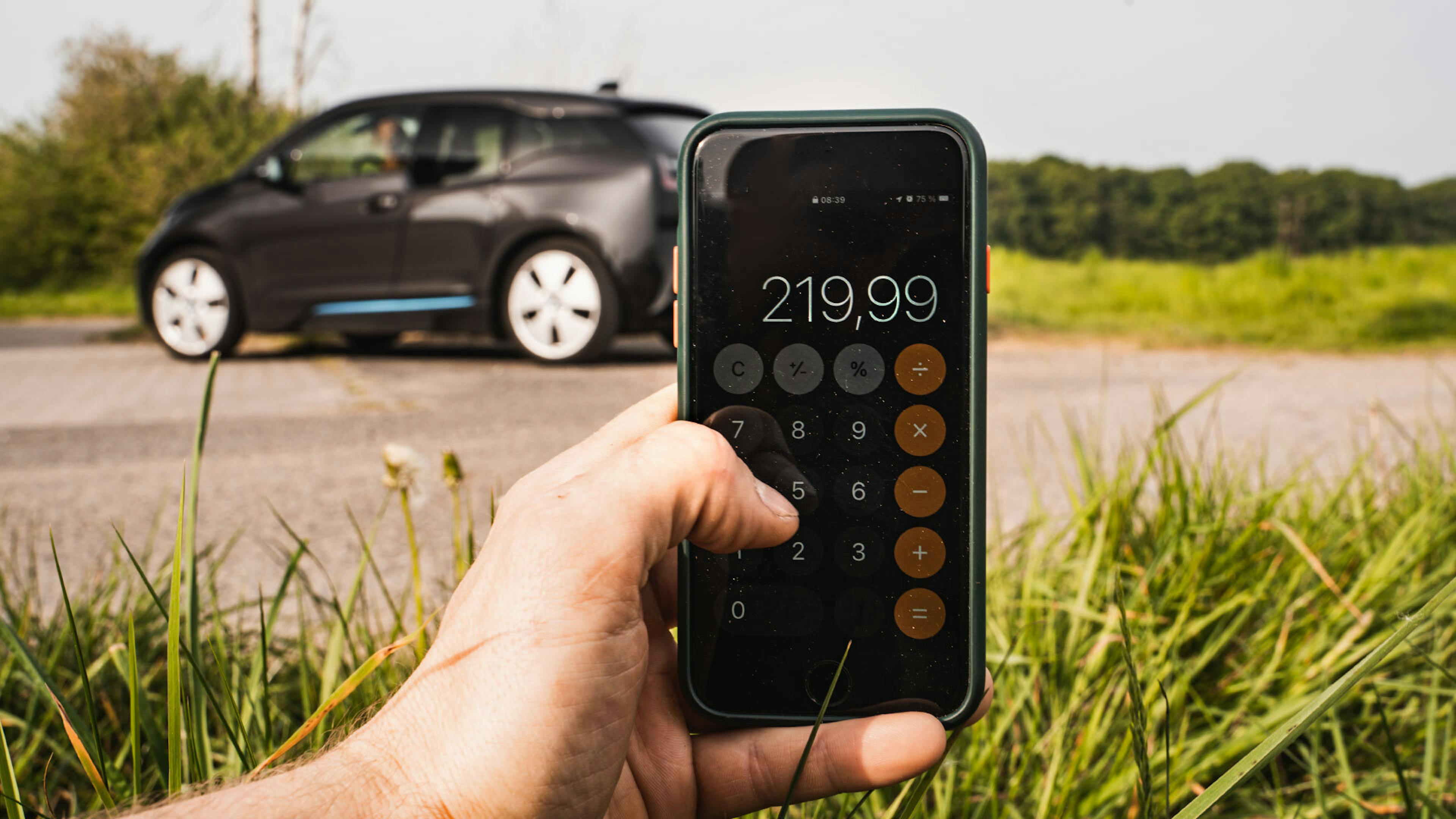 Ein Handy mit Taschenrechner ist vor einem BMW und einem Feldrand zu sehen