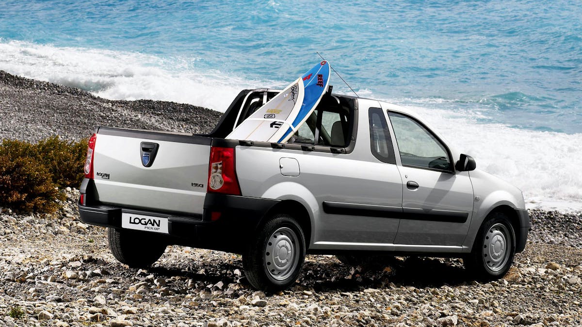 Ein silberner Dacia Logan mit einem Surfboard auf der Ladefläche steht an einem Kiesstrand.