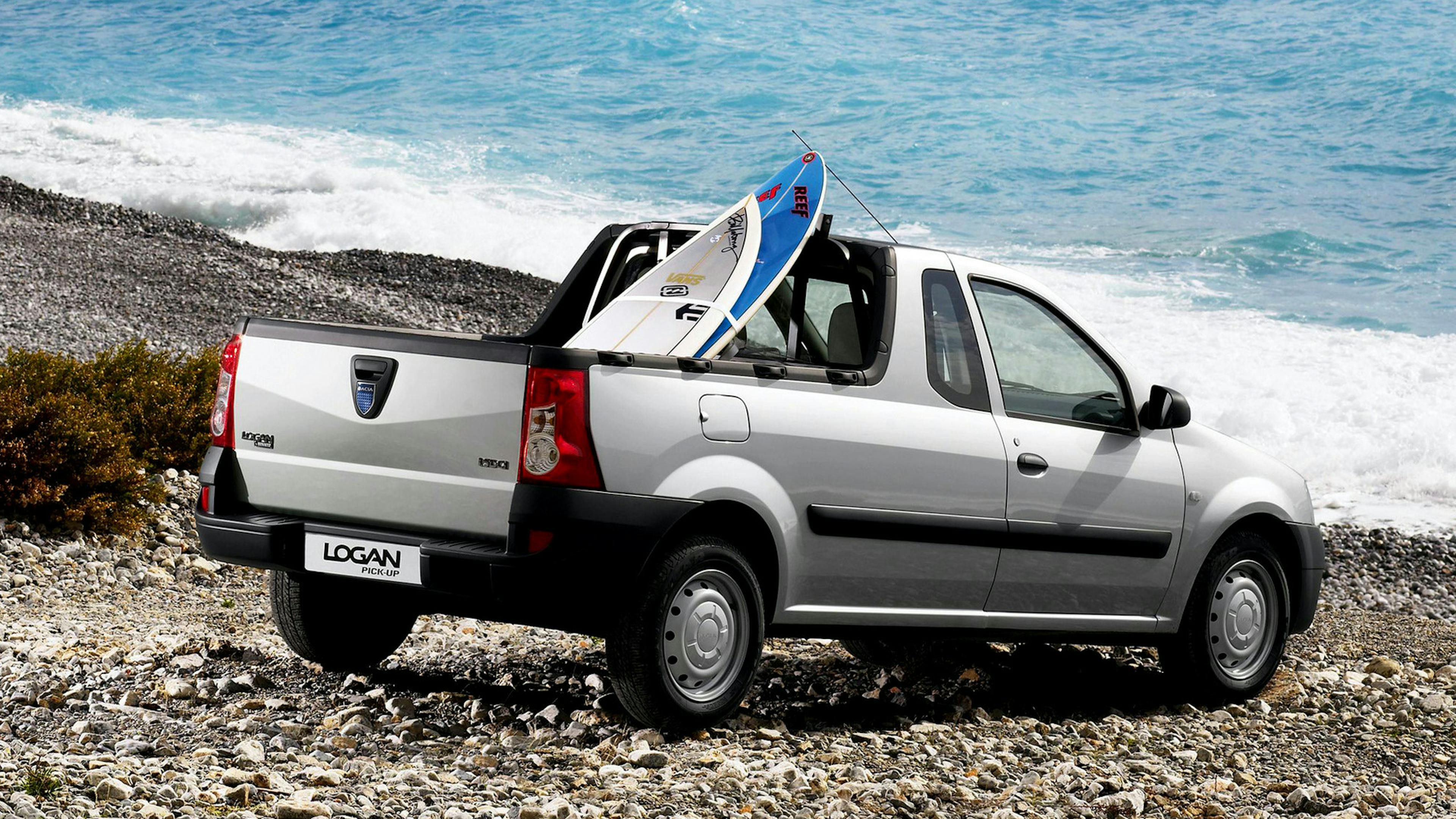 Ein silberner Dacia Logan mit einem Surfboard auf der Ladefläche steht an einem Kiesstrand.