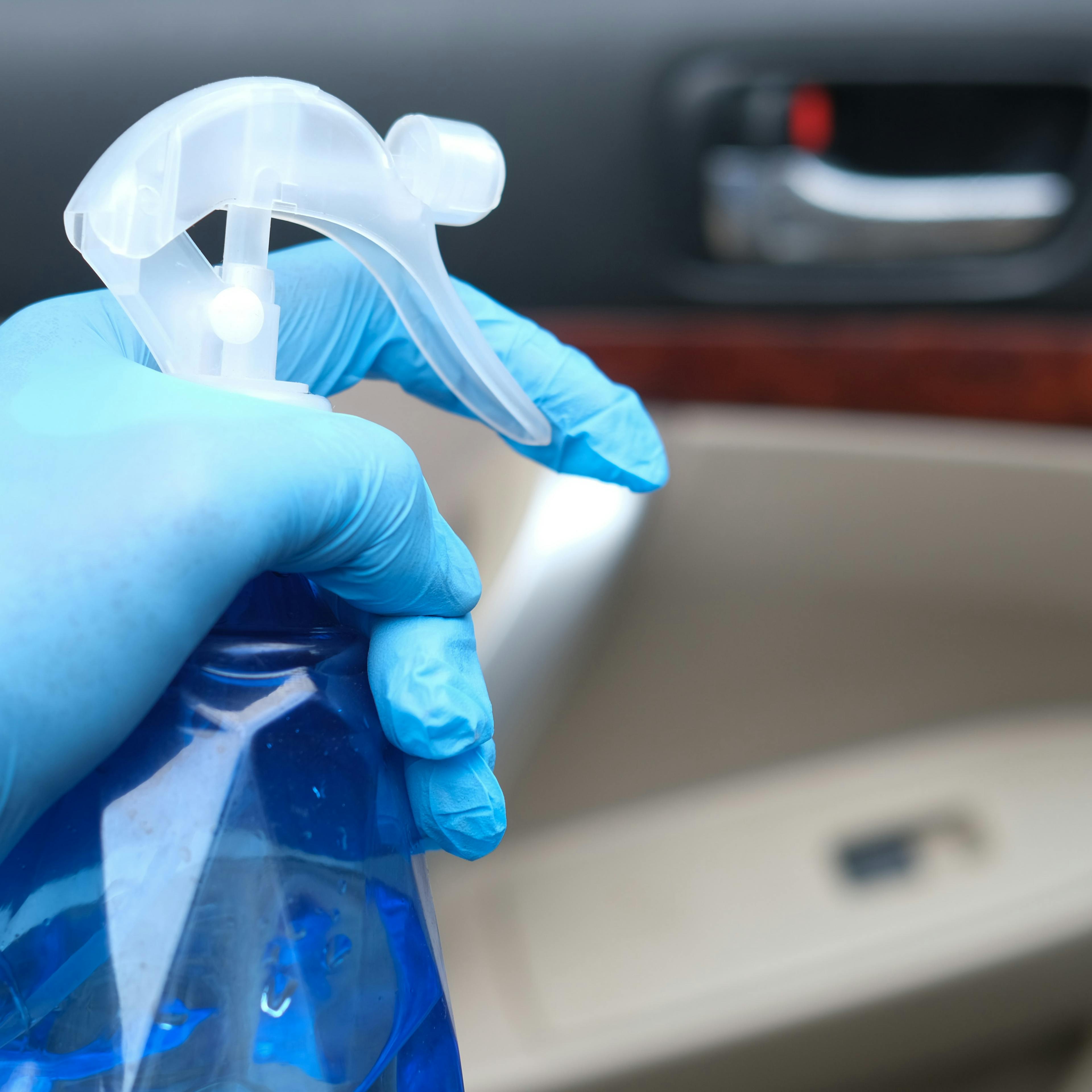 Detailaufnahme einer Hand mit blauem Handschuh und blauer Sprühflasche beim Reinigen eines Auto-Innenraumes