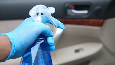 Detailaufnahme einer Hand mit blauem Handschuh und blauer Sprühflasche beim Reinigen eines Auto-Innenraumes
