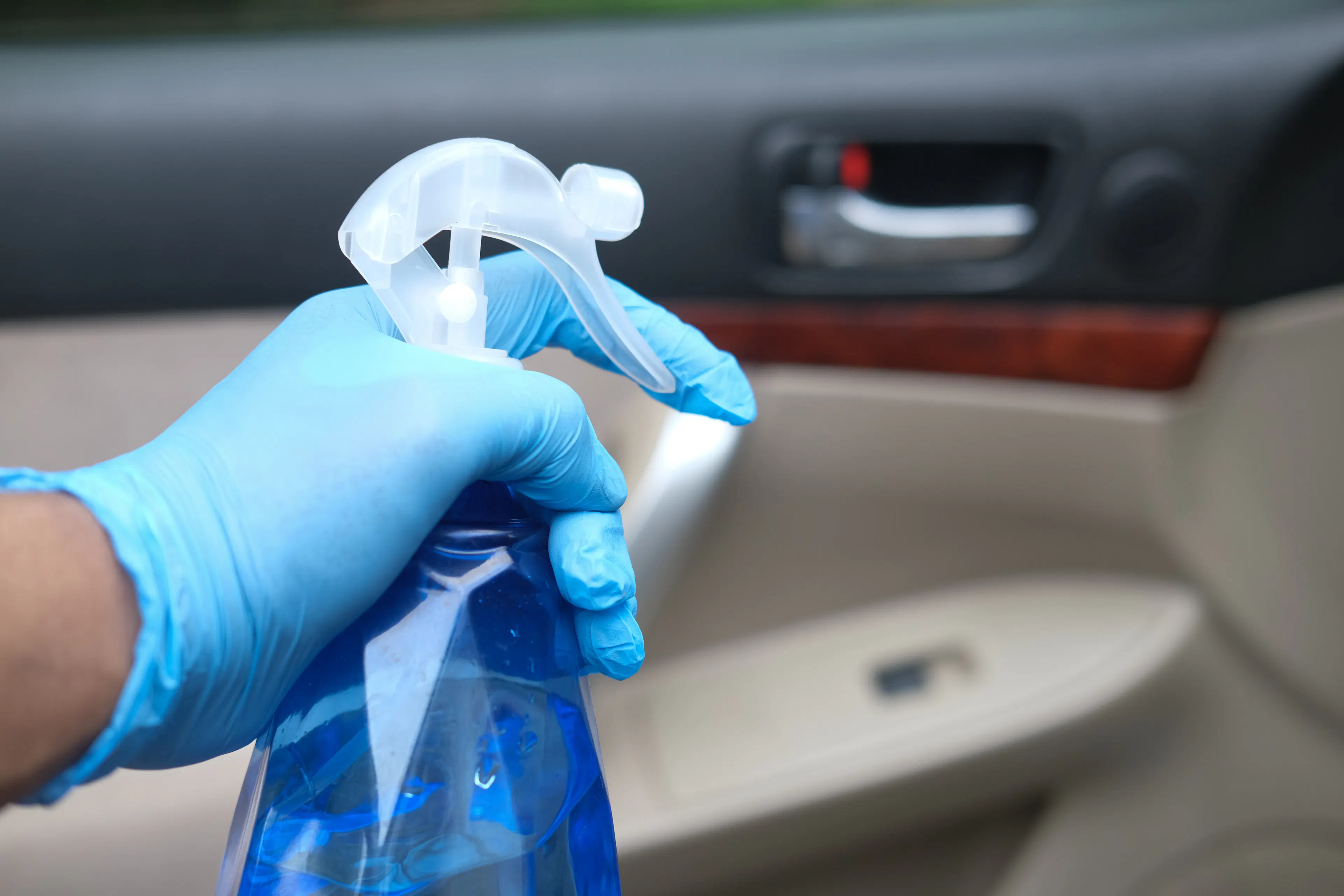 Detailaufnahme einer Hand mit blauem Handschuh und blauer Sprühflasche beim Reinigen eines Auto-Innenraumes