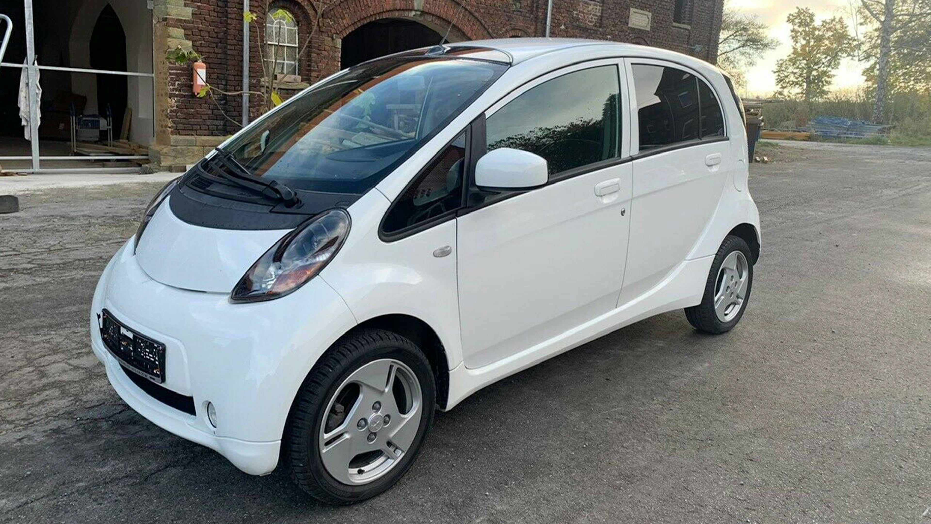 Ein weißer Mitsubishi i-MiEV steht auf einem Parkplatz vor einem Backsteingebäude.