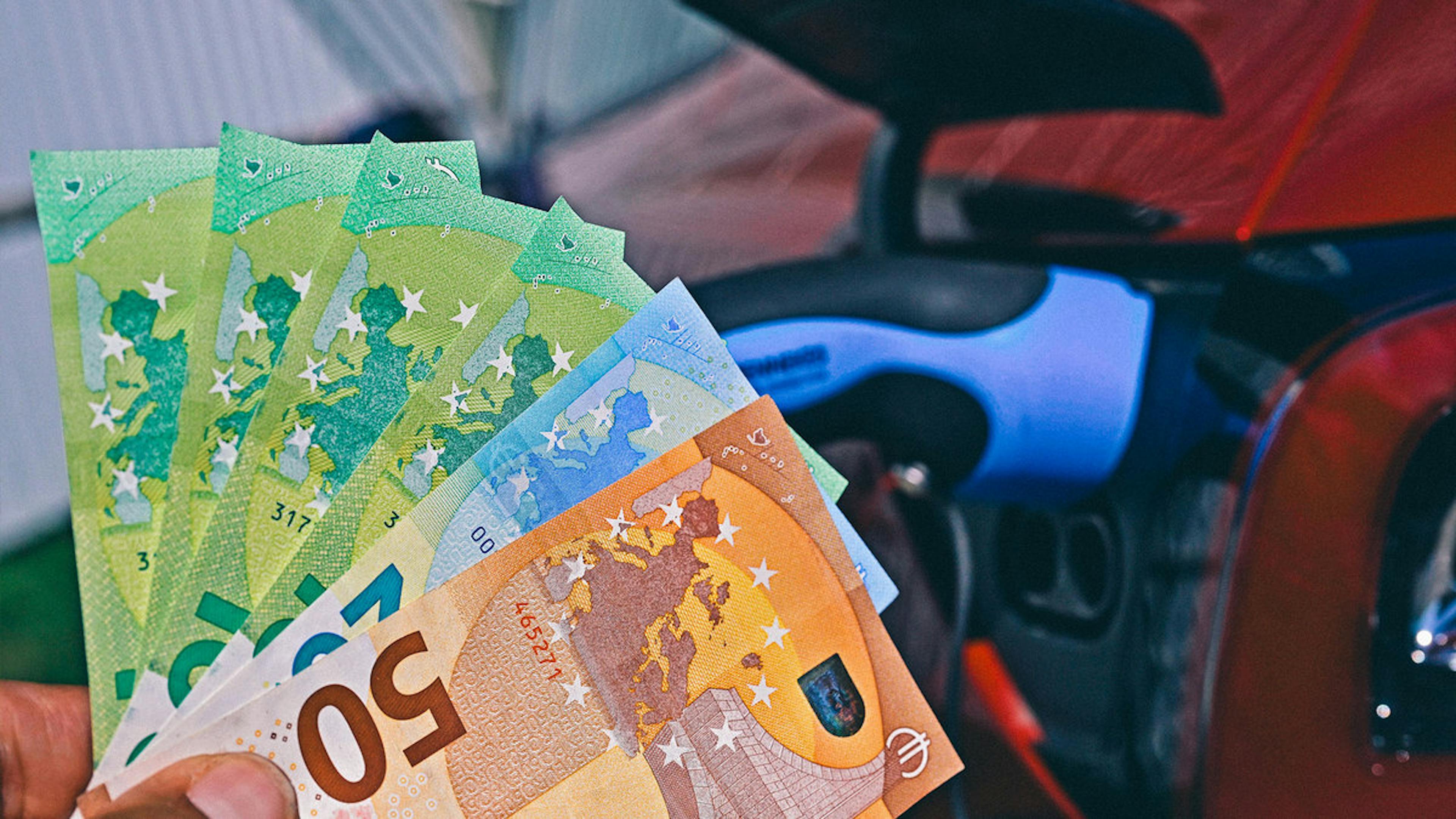 Eine Hand hält Geldscheine, im Hintergrund ist der Innenraum eines Fahrzeugs