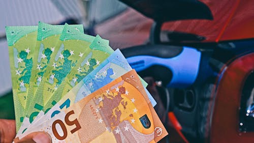 Eine Hand hält Euro-Geldscheine vor einem Elektroauto, das an einer Ladestation geladen wird.