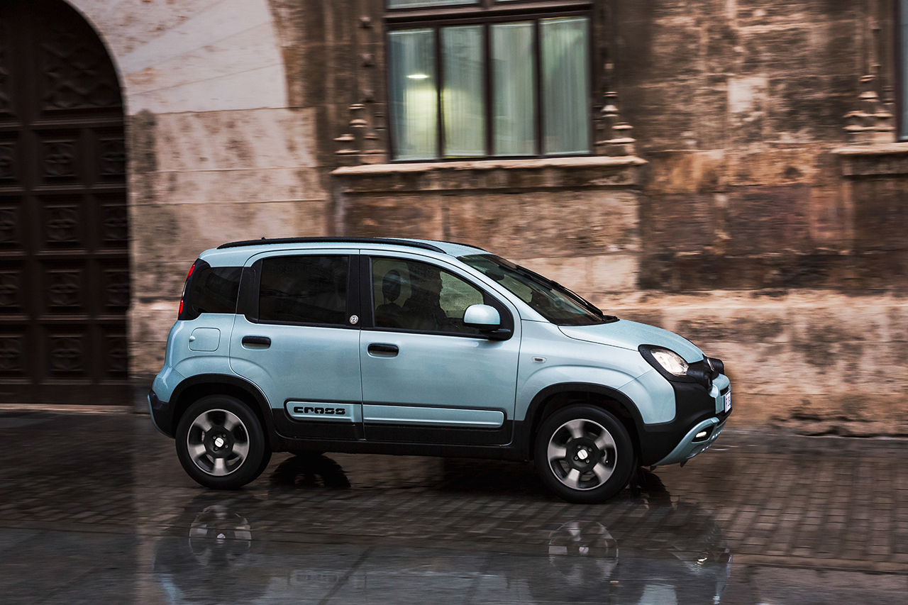 Hellblauer Fiat Panda Cross fährt auf nassem Kopfsteinpflaster an einer Steinfassade vorbei.