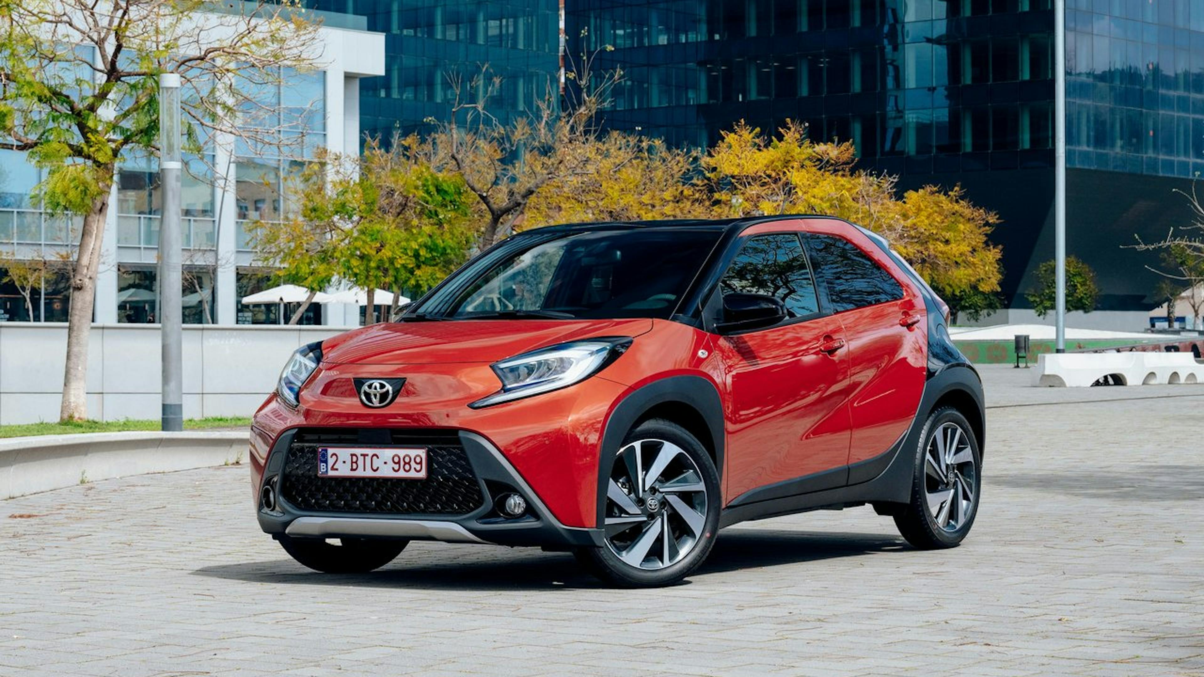 An orange Toyota Aygo X stands in front of an urban background.