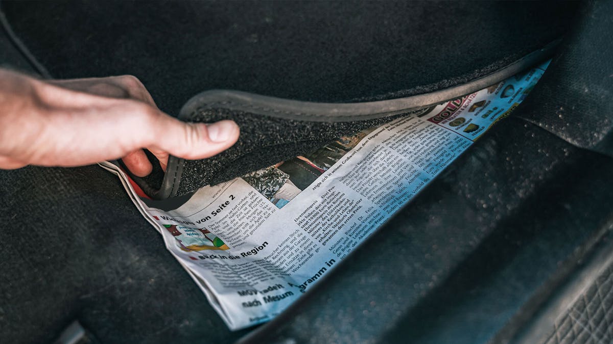 Eine Person legt ein Stück Zeitungspapier unter die Fußmatte eines Pkw.
