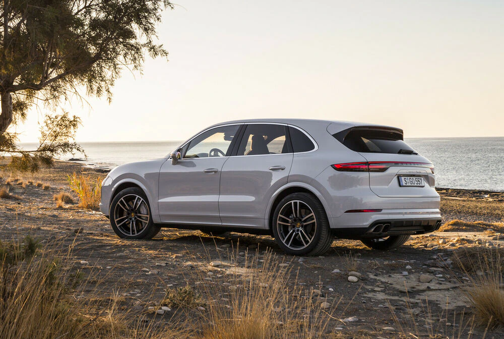 Weißes SUV parkt am Meer, umgeben von Gras und Bäumen bei Sonnenuntergang. Im Hintergrund ruhiges Wasser und klarer Himmel.