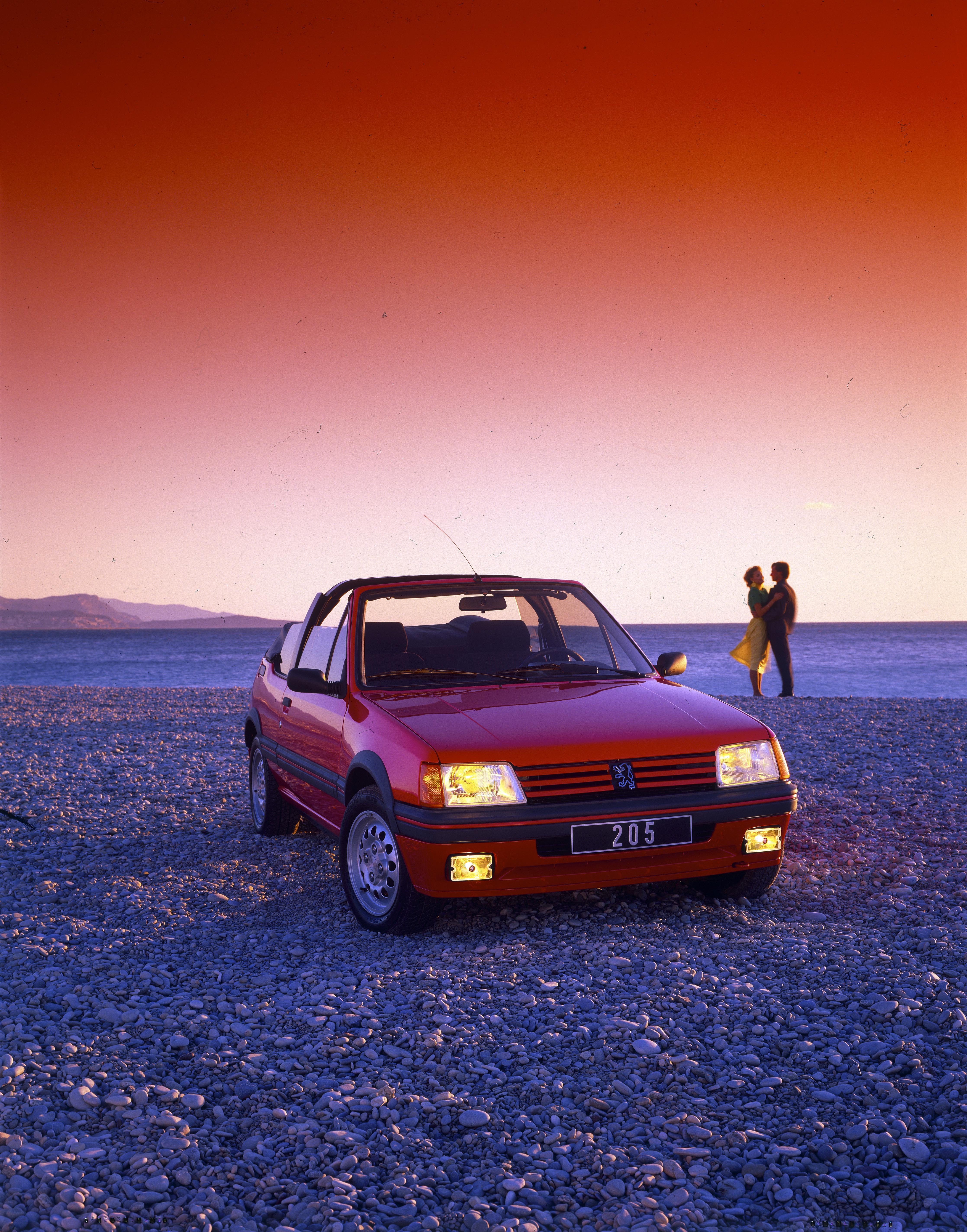 Roter Peugeot 205 Cabriolet in der Frontansicht an einem Kieselstrand bei Sonnenuntergang, zwei Personen im Hintergrund.