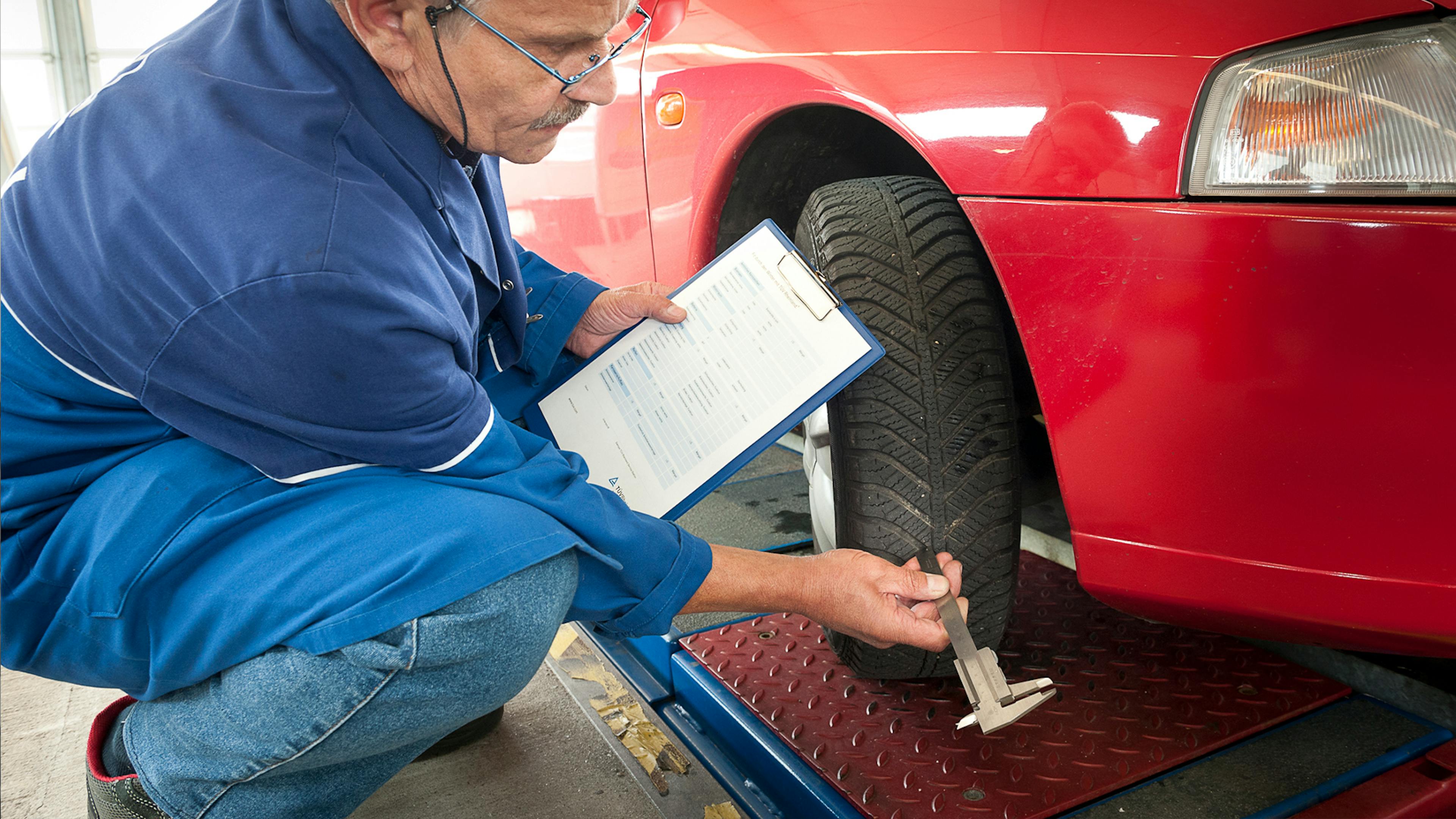 Ein Mitarbeiter des TÜV prüft die Profiltiefe eines Reifens an einem roten Pkw