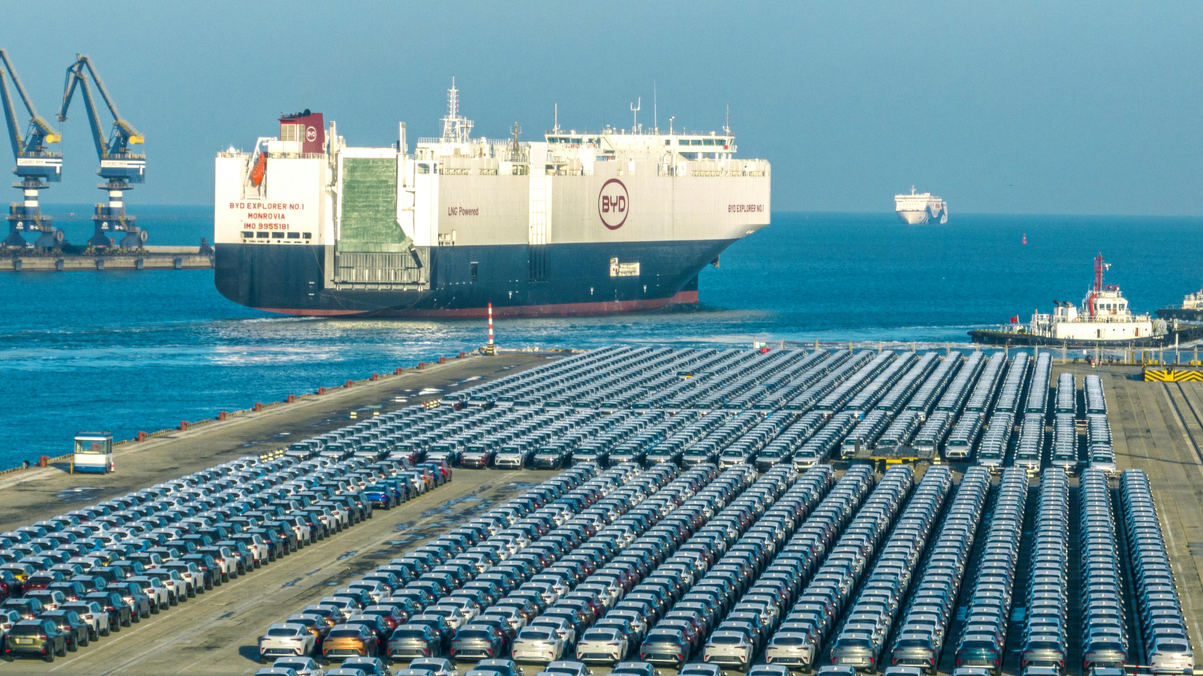 A port with new cars parked in a row with a transport ship in the background.