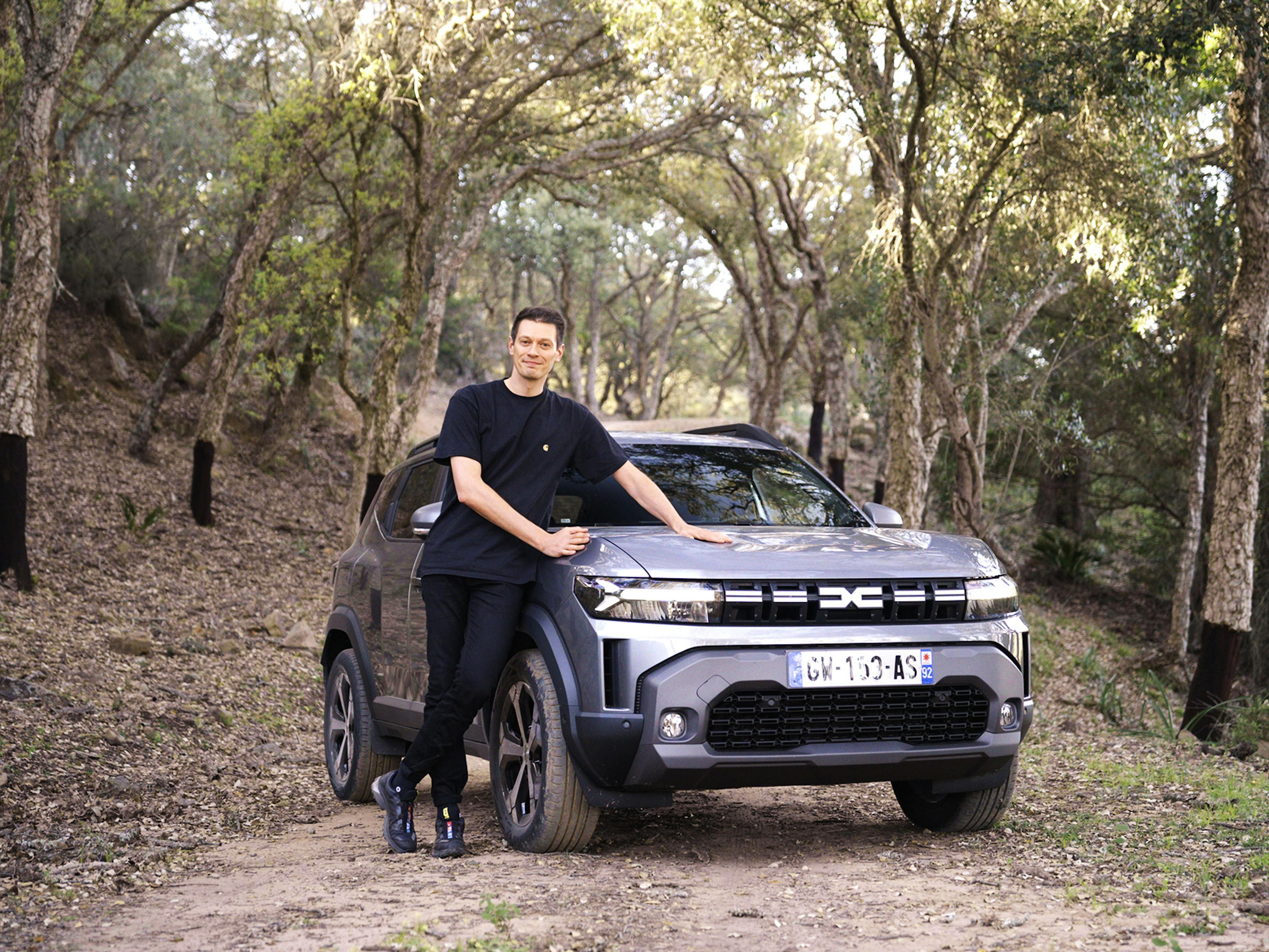 Ein junger Mann lehnt lächelnd an einem grauen SUV auf einem Waldweg, umgeben von Bäumen.