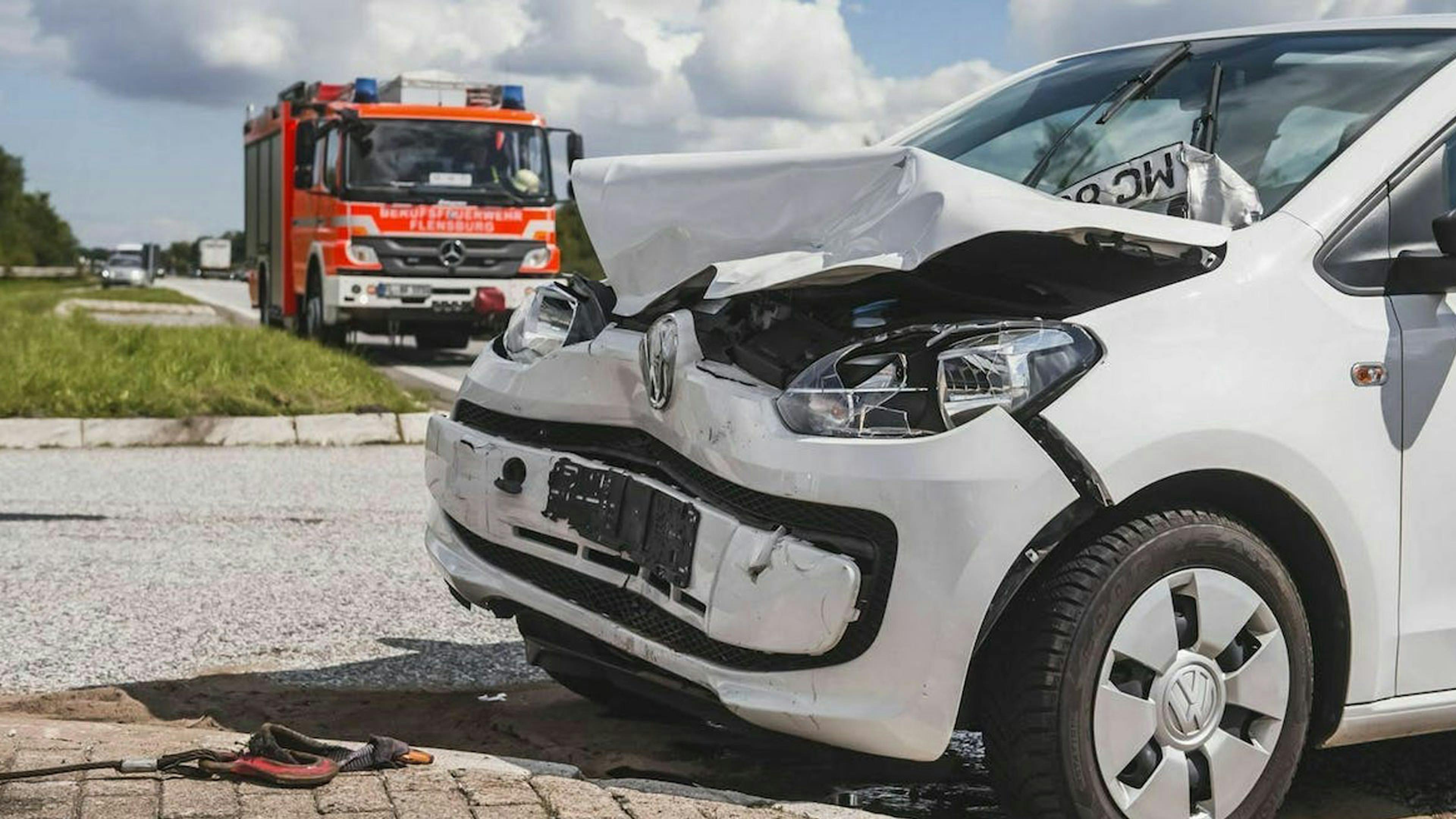 Ein verunfallter weißer Pkw steht am Rand einer Straße, im Hintergrund steht ein Feuerwehrfahrzeug.