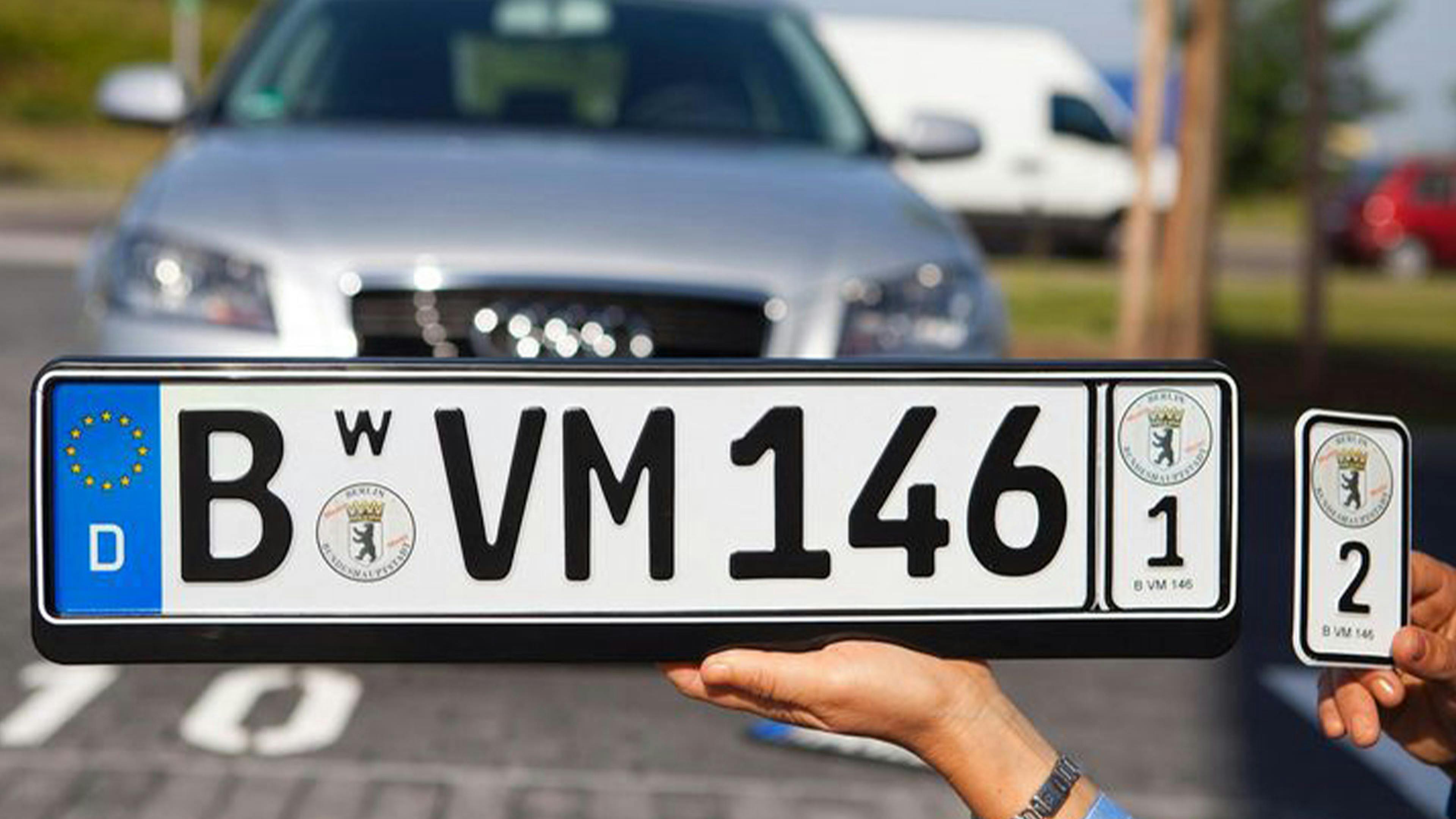Nahaufnahme eines deutschen Kfz-Kennzeichens mit Berliner Wappen, gehalten vor einem geparkten Audi. Eine Hand hält zusätzlich ein kleines Schild.