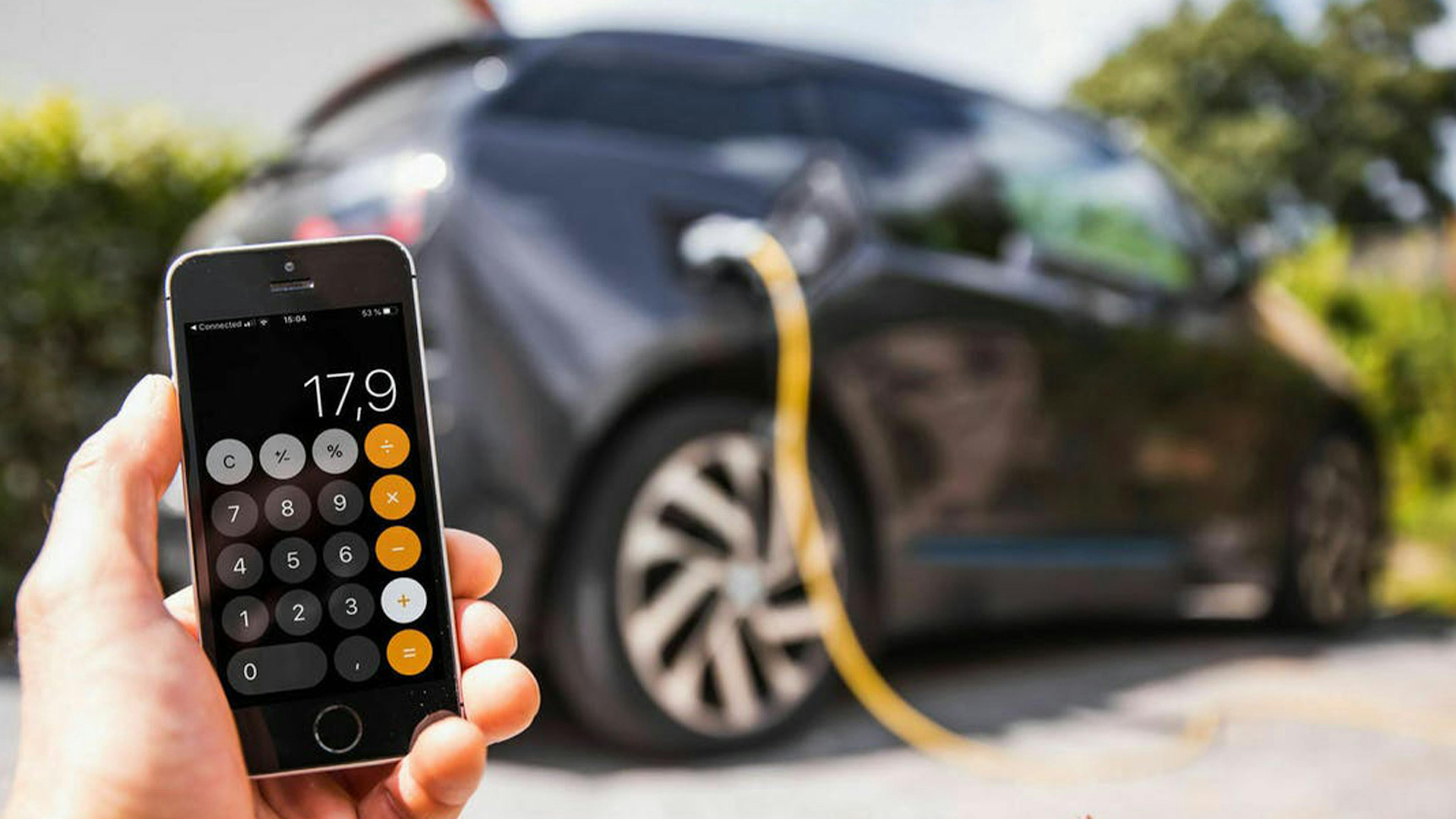 Eine Hand hält ein Smartphone, auf dem ein Taschenrechner aktiviert ist, im Hintergrund lädt ein Elektroauto.