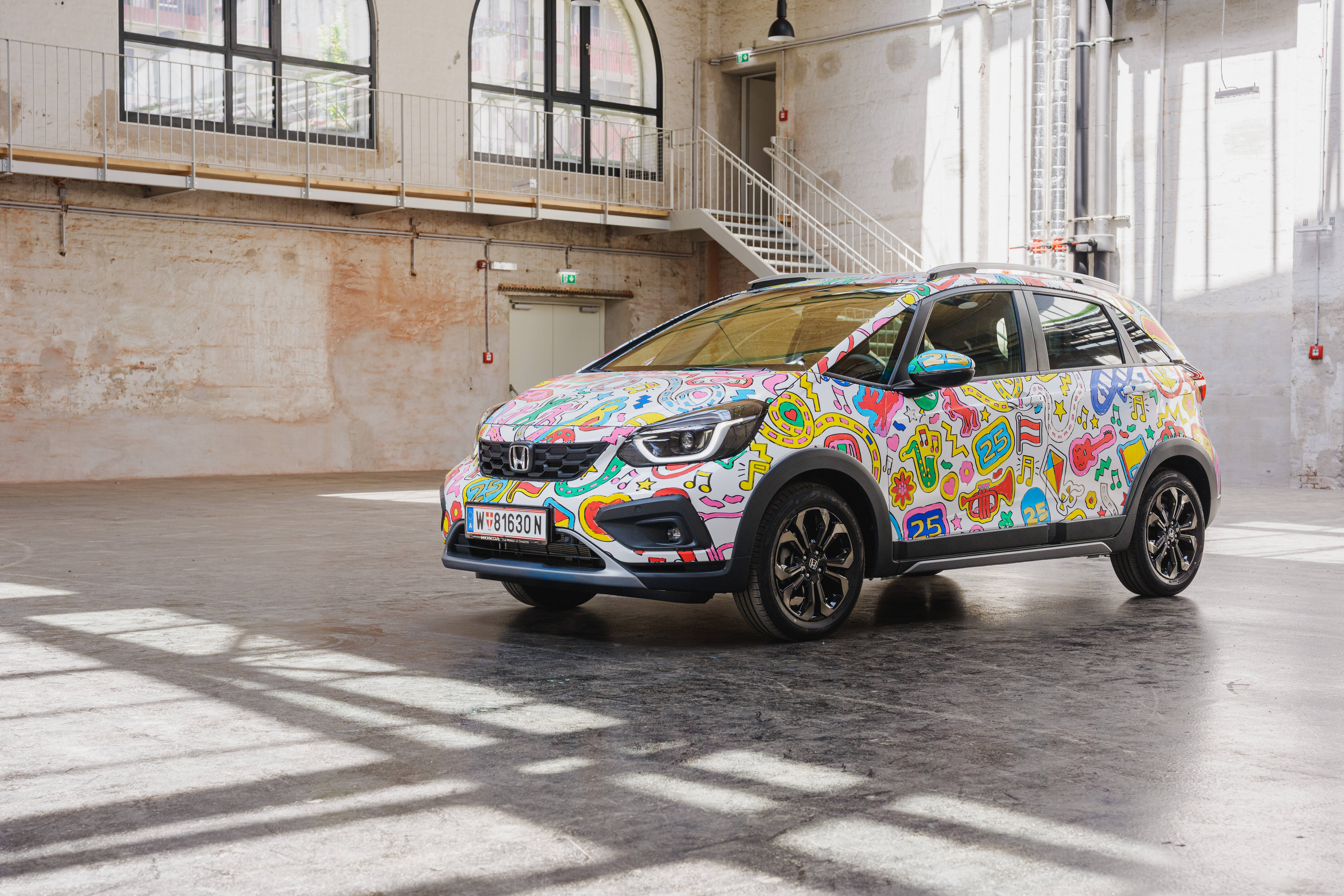 Honda Jazz mit bunter Folierung aus Musikinstrumenten und der Zahl 25 in einer Industriehalle mit Backsteinwand.