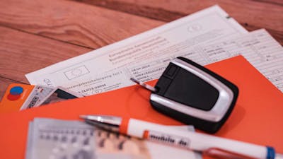 Eine orangefarbene Mappe, ein Autokaufvertrag, Bargeld, Kreditkarten, ein Autoschlüssel und ein Kugelschreiber liegen auf einem Holztisch.