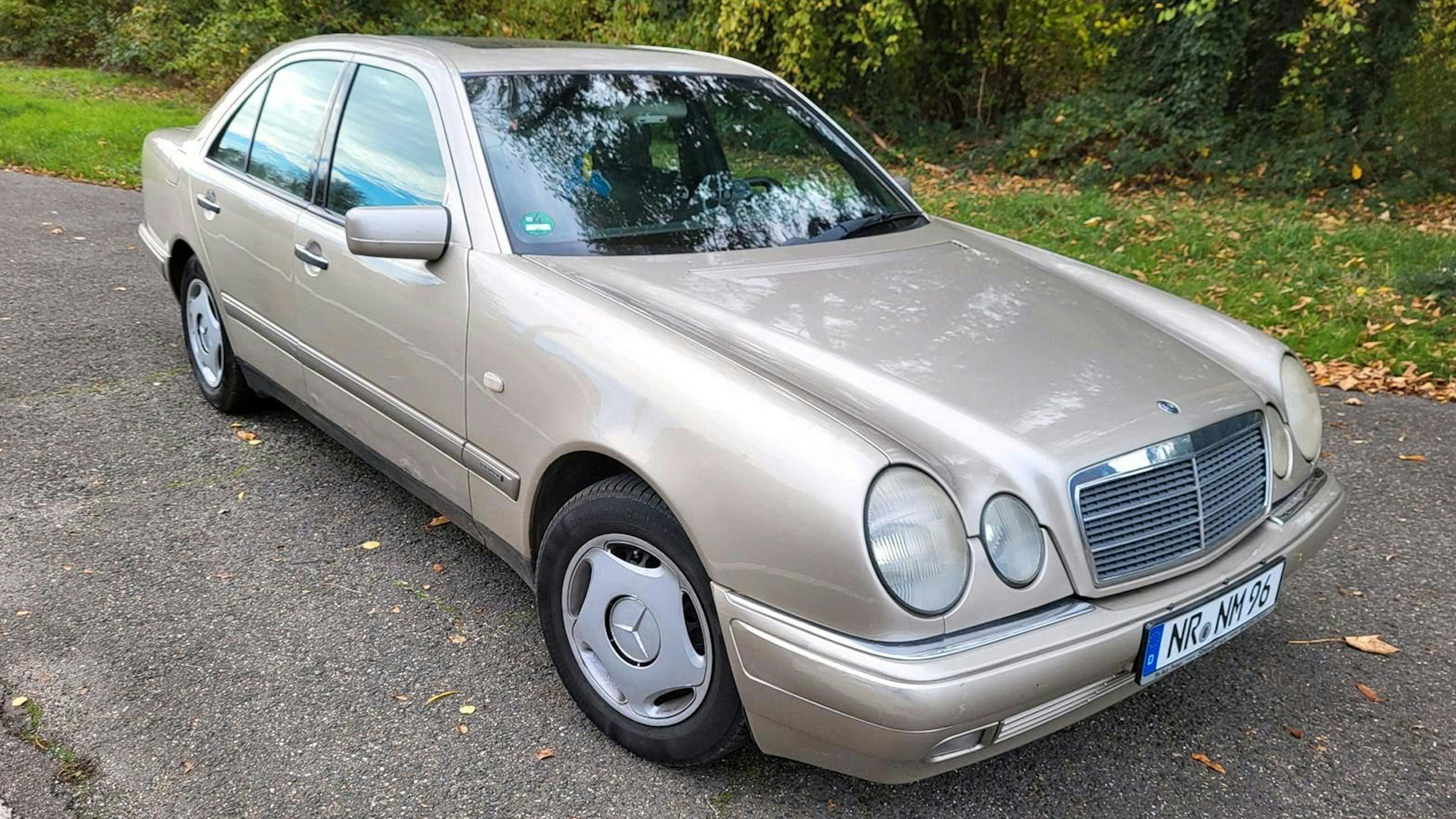 Eine silberne Mercedes-Benz E-Klasse steht an einem Straßenrand mit Grünflächen im Hintergrund.