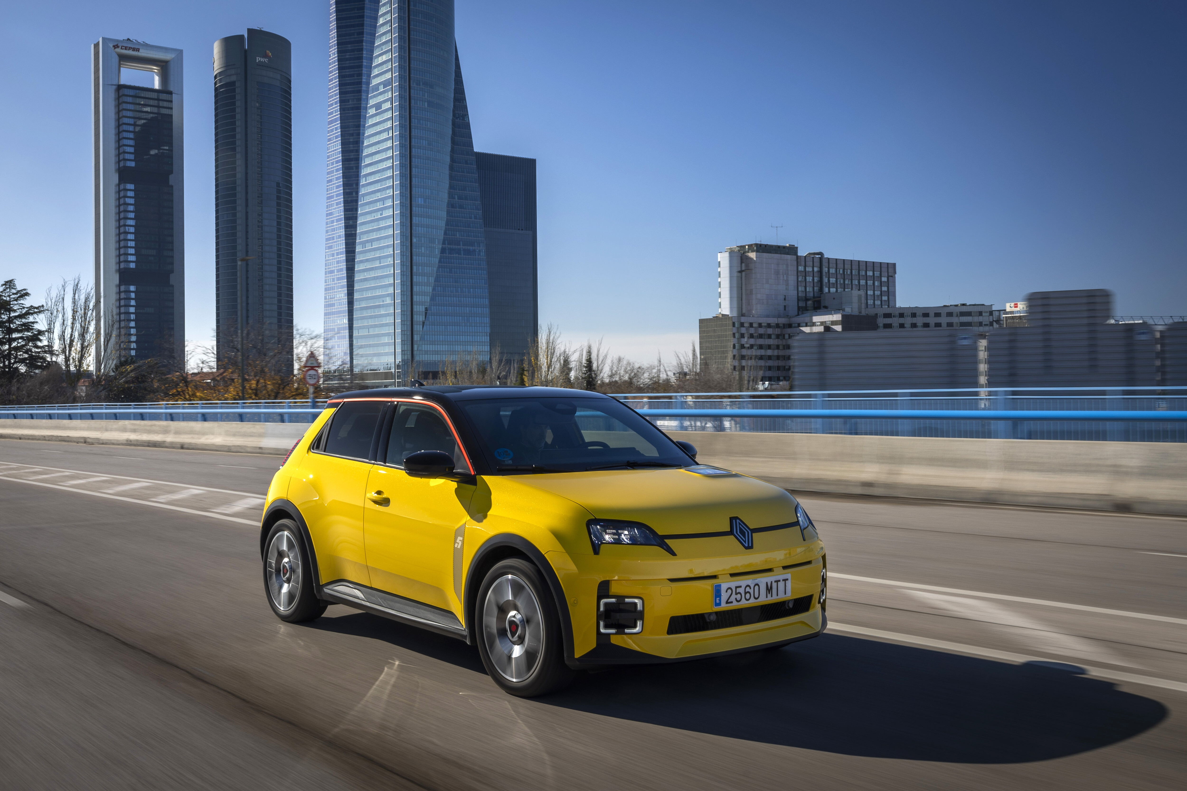 Gelber Renault 5 E-Tech mit schwarzem Dach fährt auf einer Straße, im Hintergrund ragen Hochhäuser in den blauen Himmel.