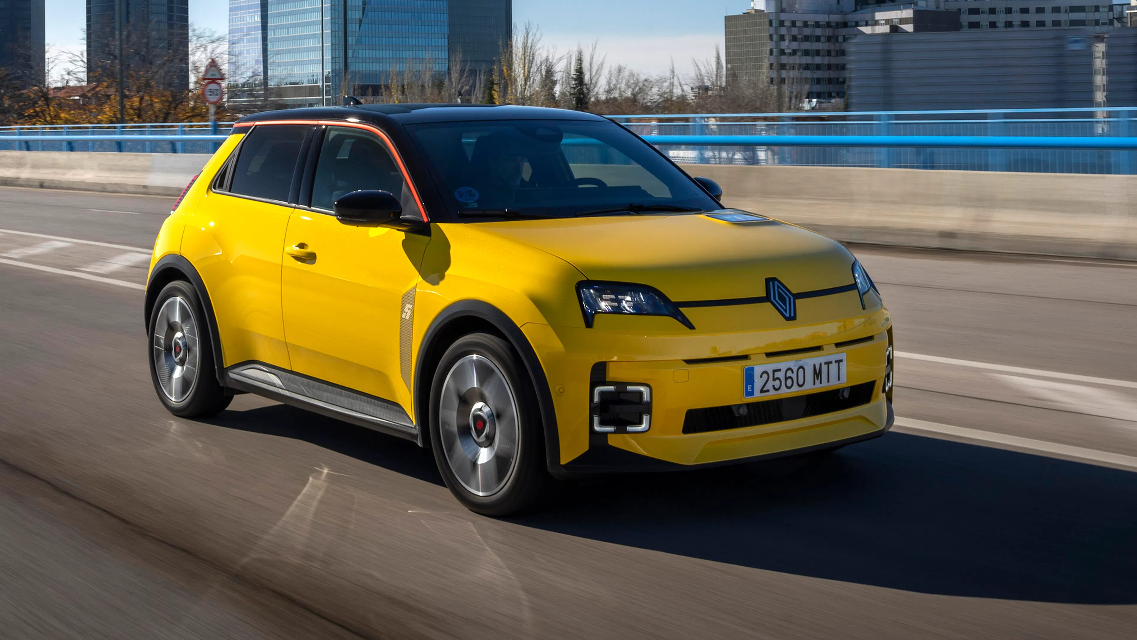 Gelber Renault 5 E-Tech mit schwarzem Dach fährt auf einer Straße, im Hintergrund ragen Hochhäuser in den blauen Himmel.