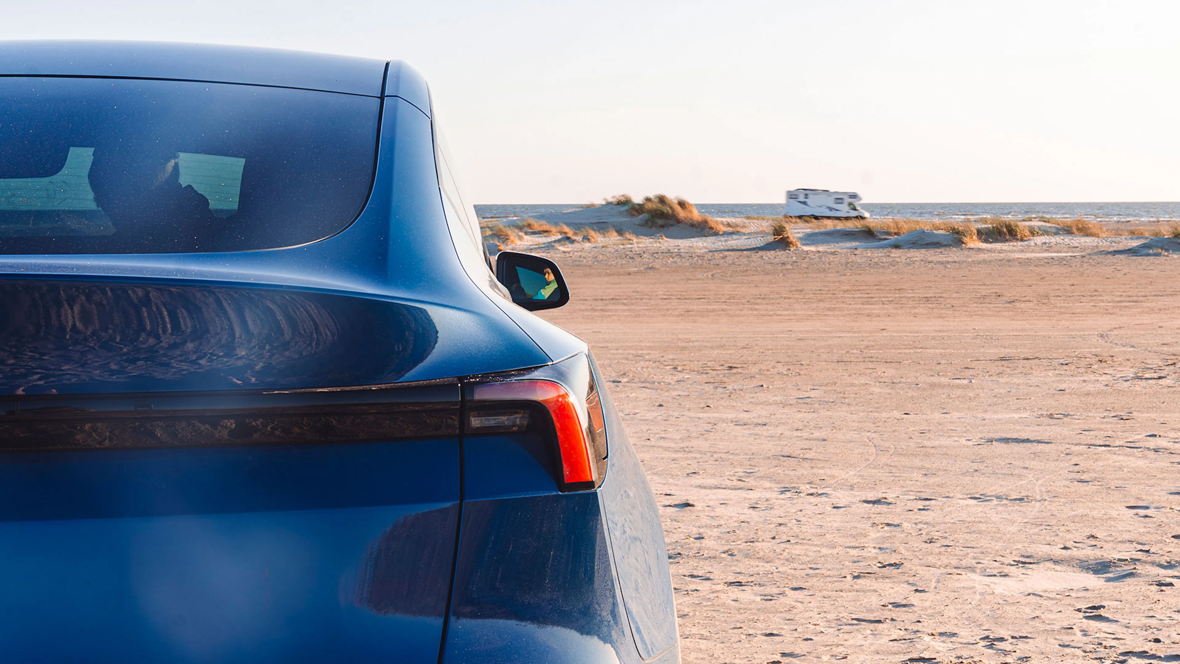 Nahaufnahme der Rückleuchte eines blaues Tesla Model Y. Im Hintergrund fährt am Strand ein Wohnmobil entlang.