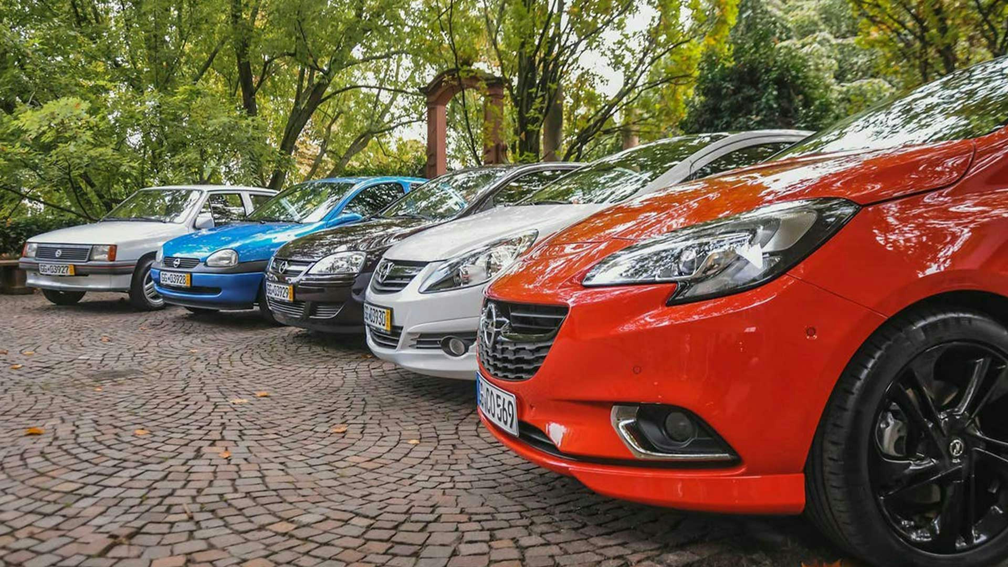 Fünf Pkw der Marke Opel stehen nebeneinander auf einem Parkplatz mit Bäumen im Hintergrund