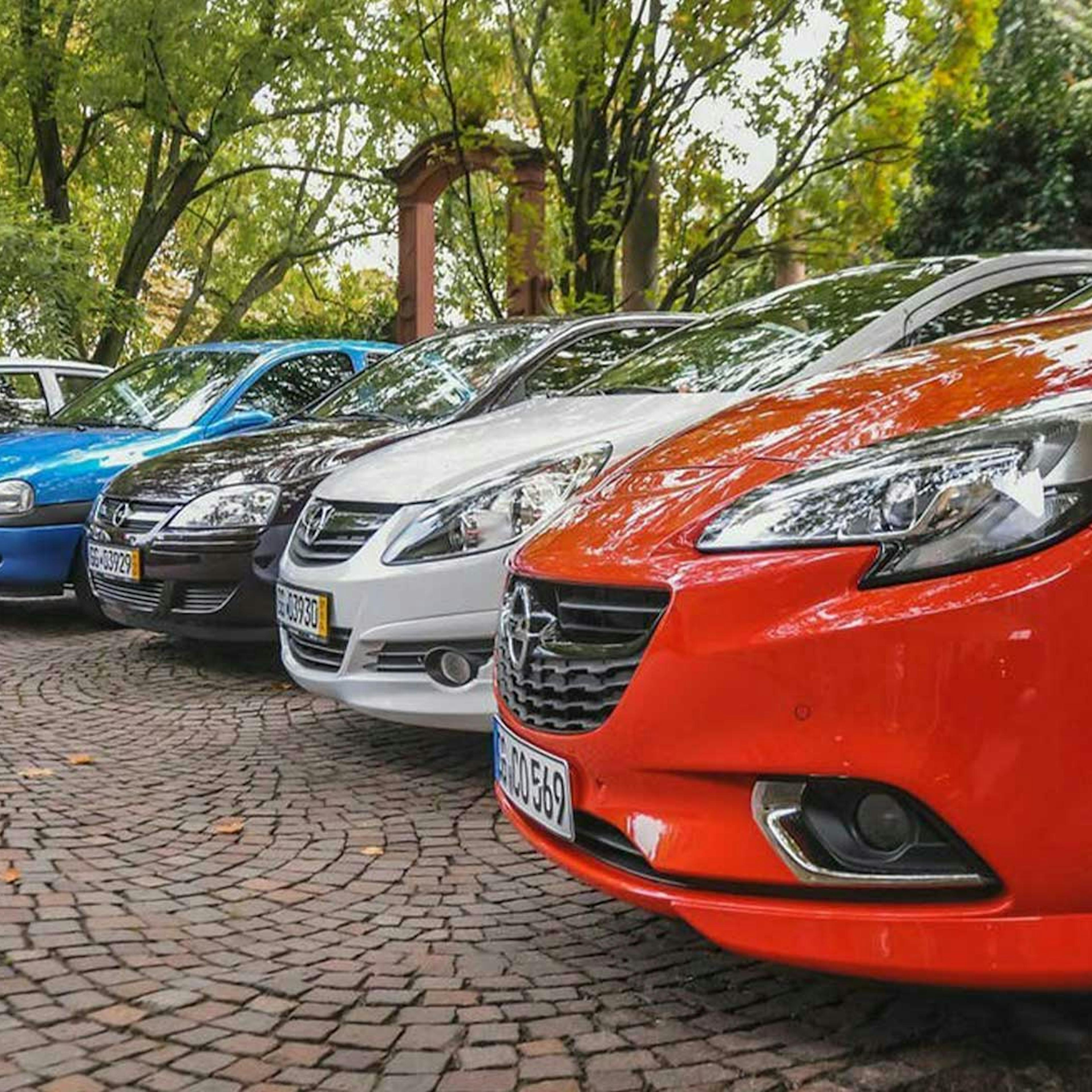 Fünf Pkw der Marke Opel stehen nebeneinander auf einem Parkplatz mit Bäumen im Hintergrund