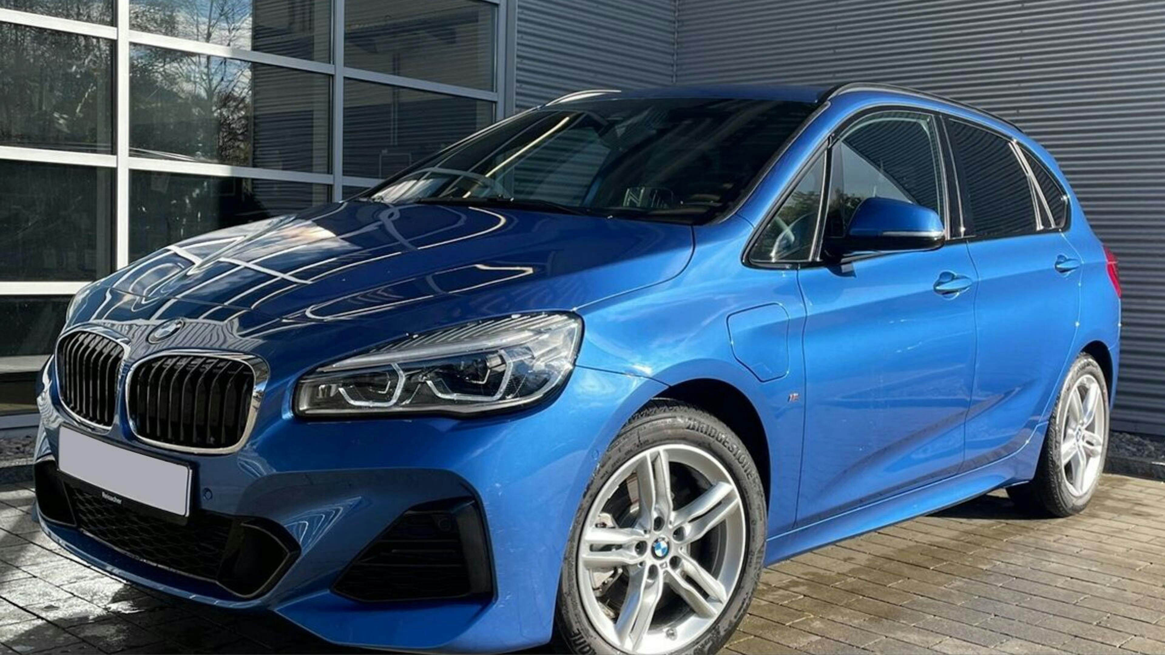 Ein blauer BMW Zweier Active Tourer Plug-in-Hybrid steht vor einem Gebäude mit tiefen Fenstern und grauem Rolltor