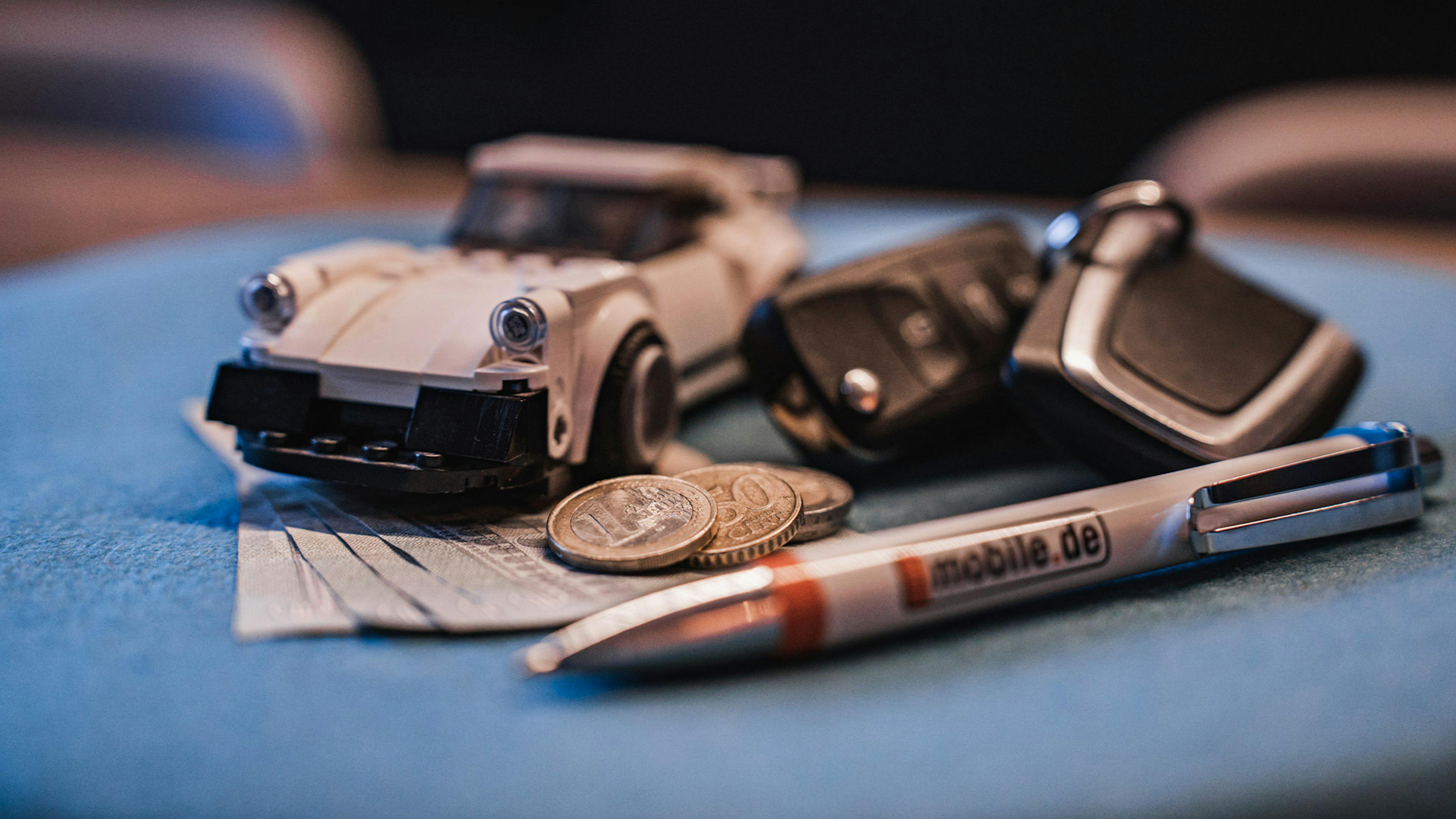 Arrangement mit einem Spielzeugauto, einem Autoschlüssel, einem Kugelschreiber und Bargeld auf blauem Untergrund.