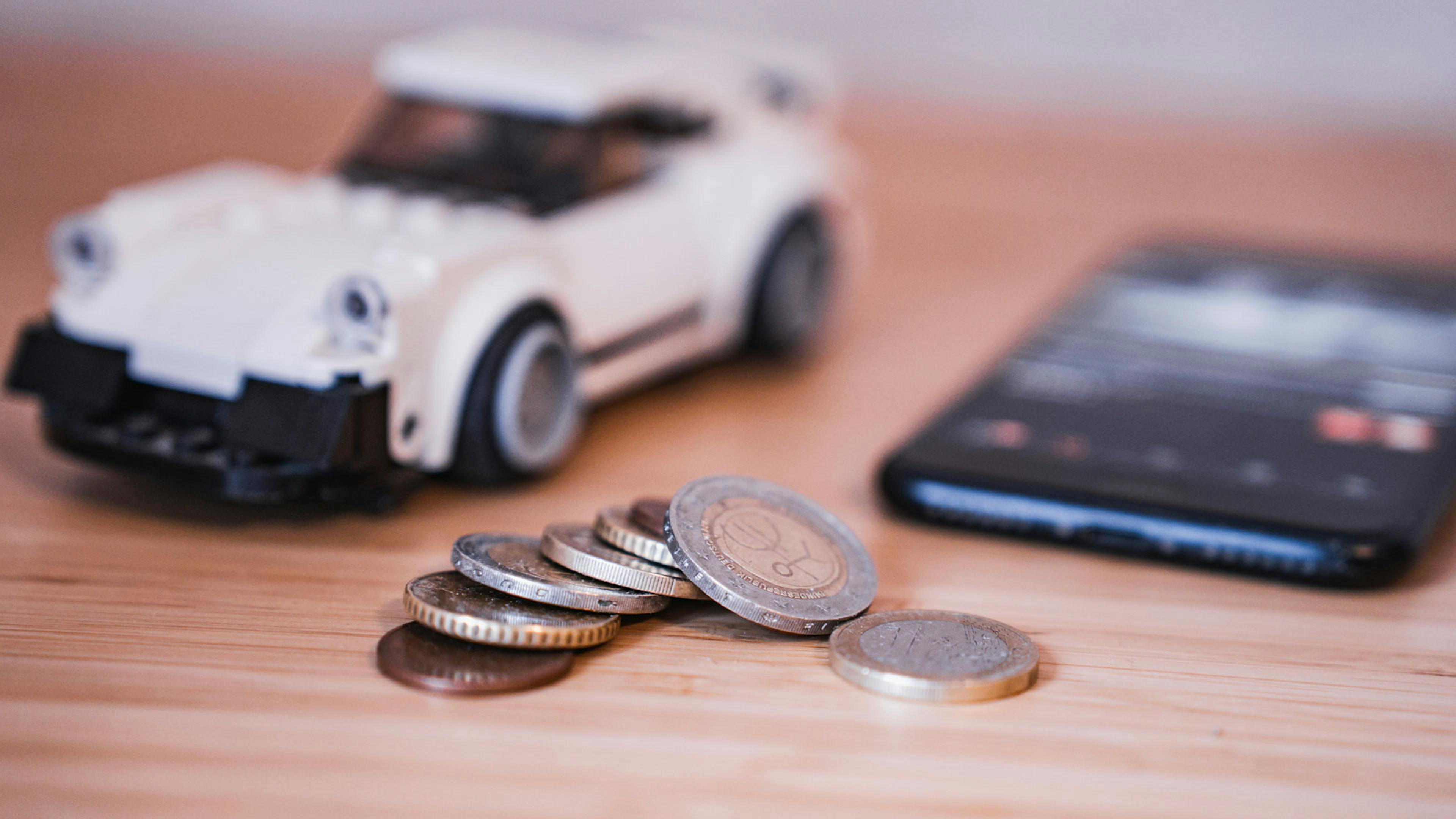 Stillleben mit einem weißen Spielzeugauto, verschiedenen Euromünzen und einem Smartphone.