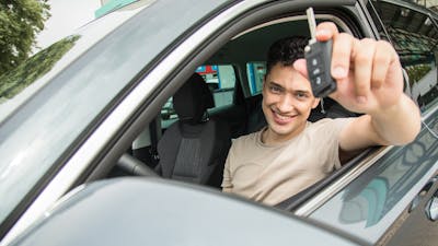 Ein junger Mann sitzt im Fahrersitz eines schwarzen Pkw und hält einen Autoschlüssel hoch.