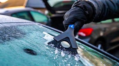 Nahaufnahme einer behandschuhten Hand, die mit einem Eiskratzer die Frontscheibe eines Autos enteist.