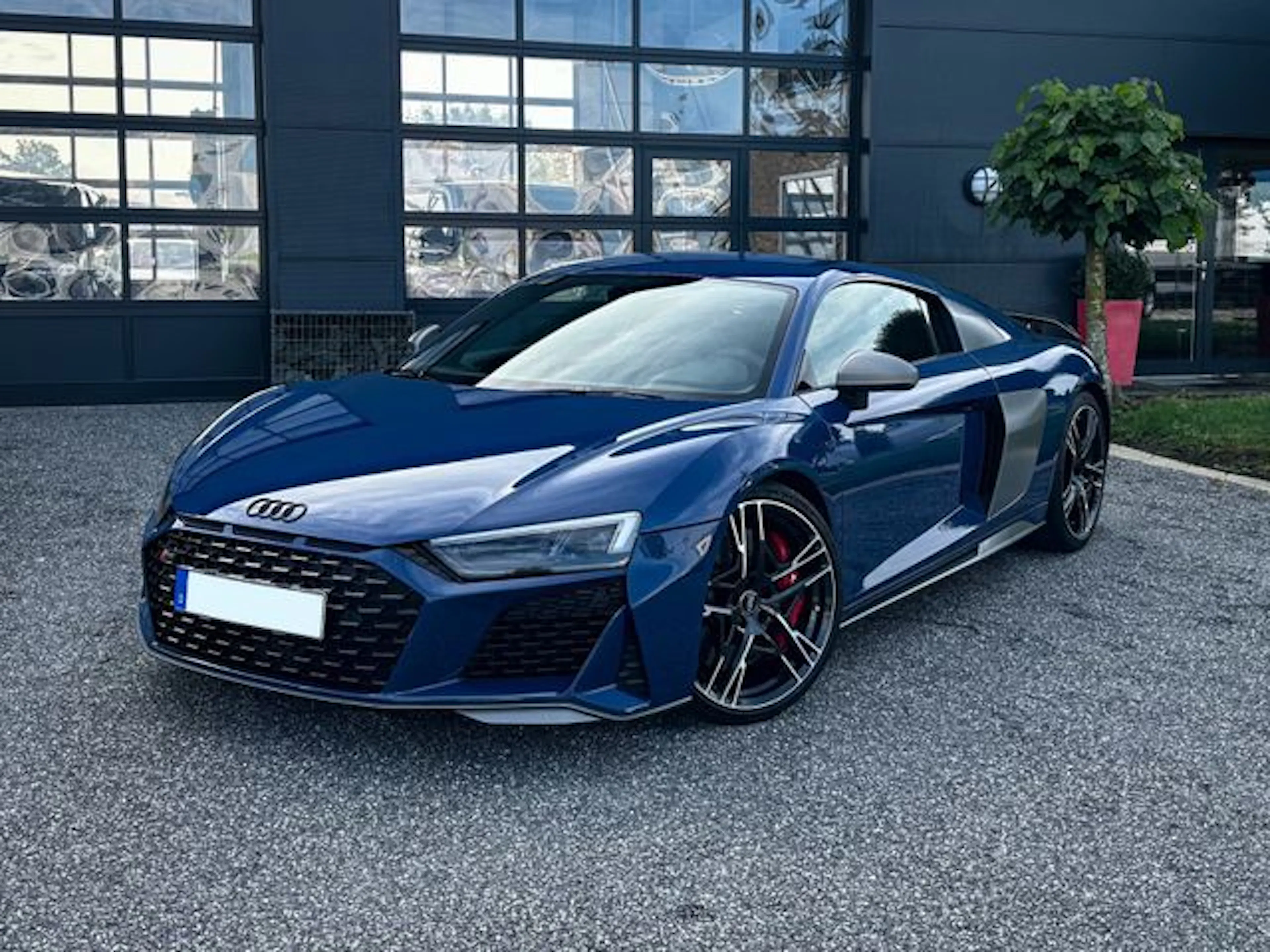 Blauer Audi R8 Sportwagen steht auf Asphalt vor einem modernen Gebäude mit Glasfassade und Pflanze im Topf.