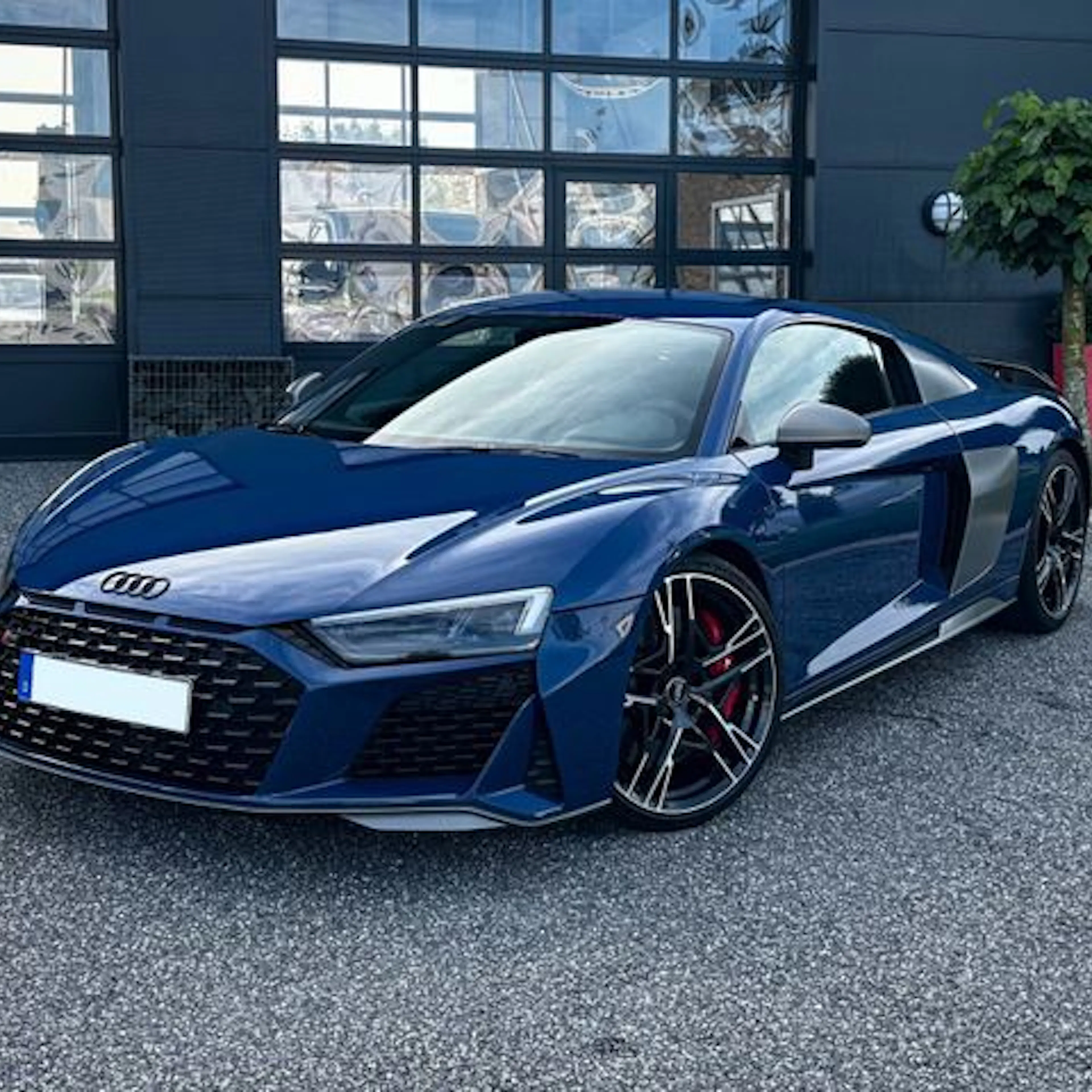 Blauer Audi R8 Sportwagen steht auf Asphalt vor einem modernen Gebäude mit Glasfassade und Pflanze im Topf.