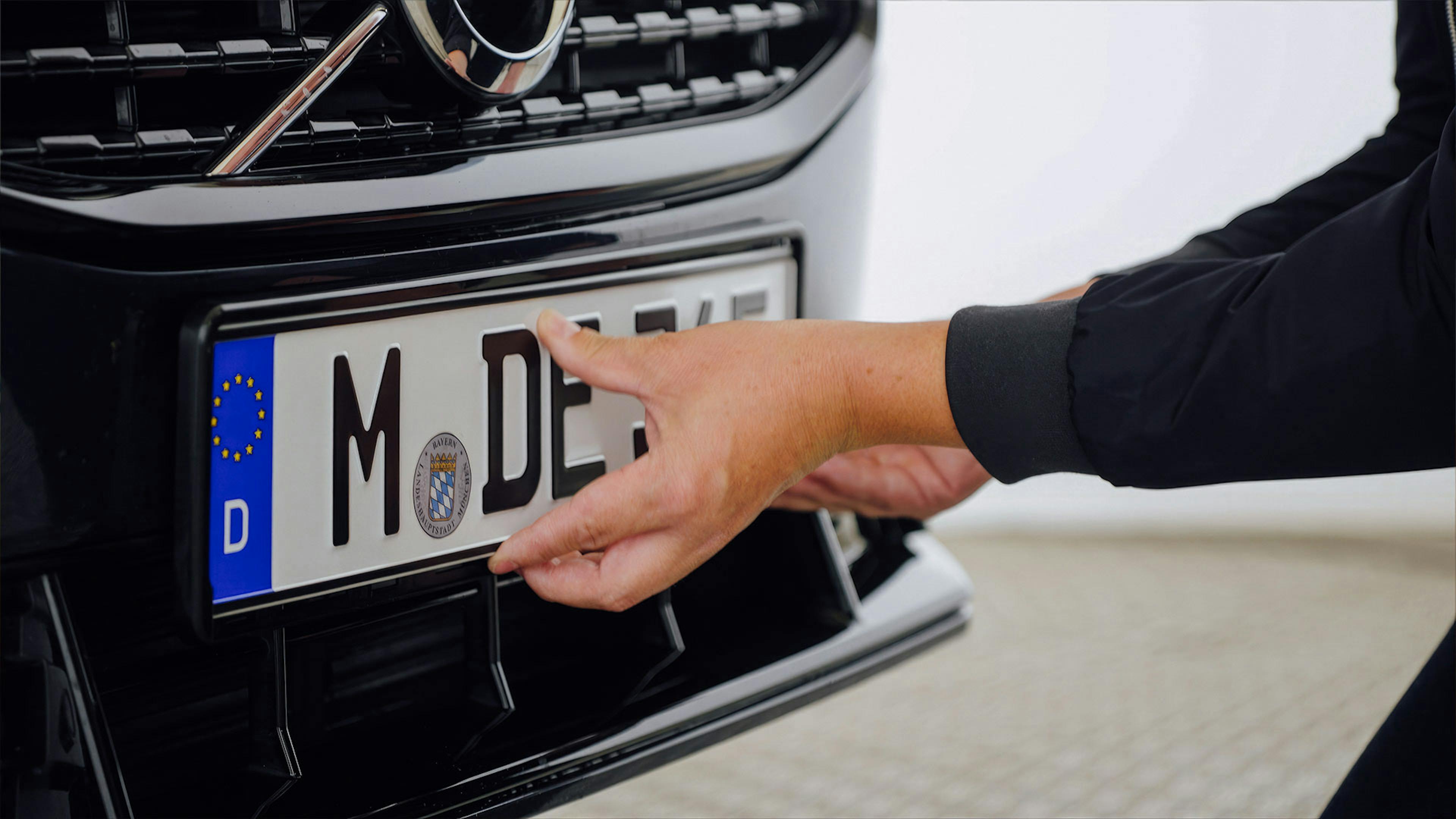 Eine Person befestigt ein deutsches Kfz-Kennzeichen an der Front eines schwarzen Autos.