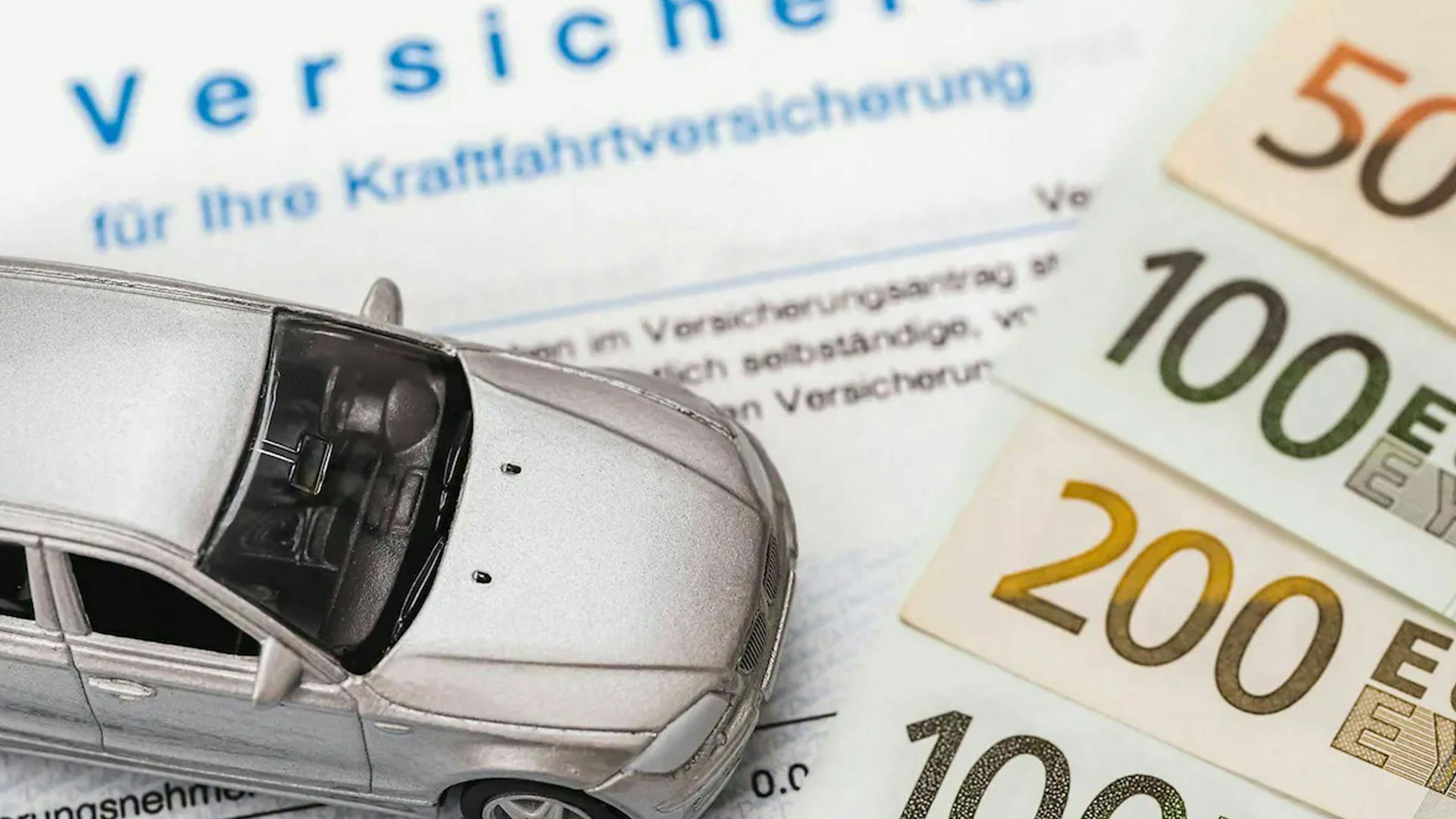 Stillleben mit einem ausgedruckten Kfz-Versicherungsvertrag, Geldscheinen und einem Spielzeugauto. 