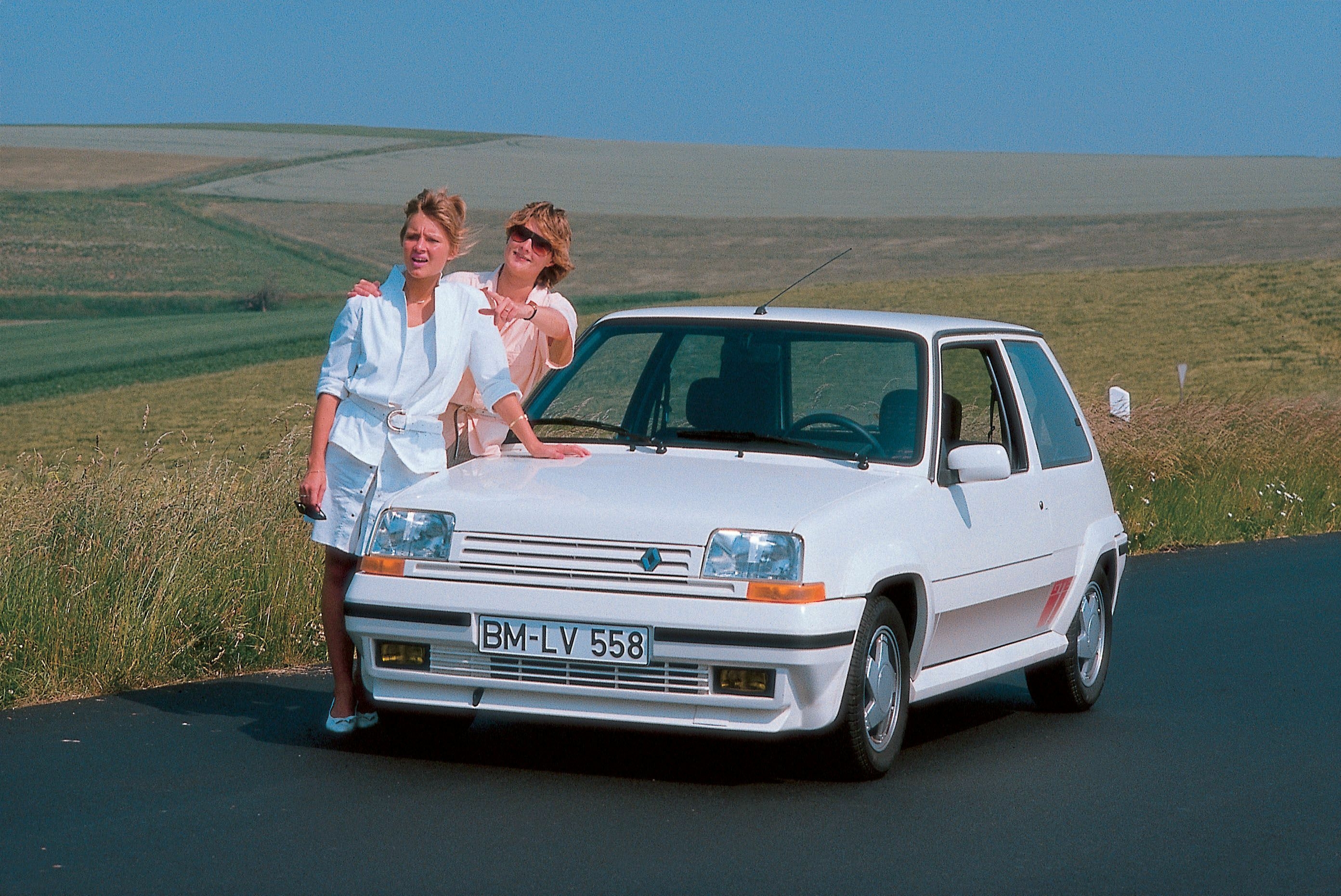 Weißer Renault 5 GT Turbo Phase 2 steht frontal auf einer Straße, zwei Personen lehnen an der Motorhaube.