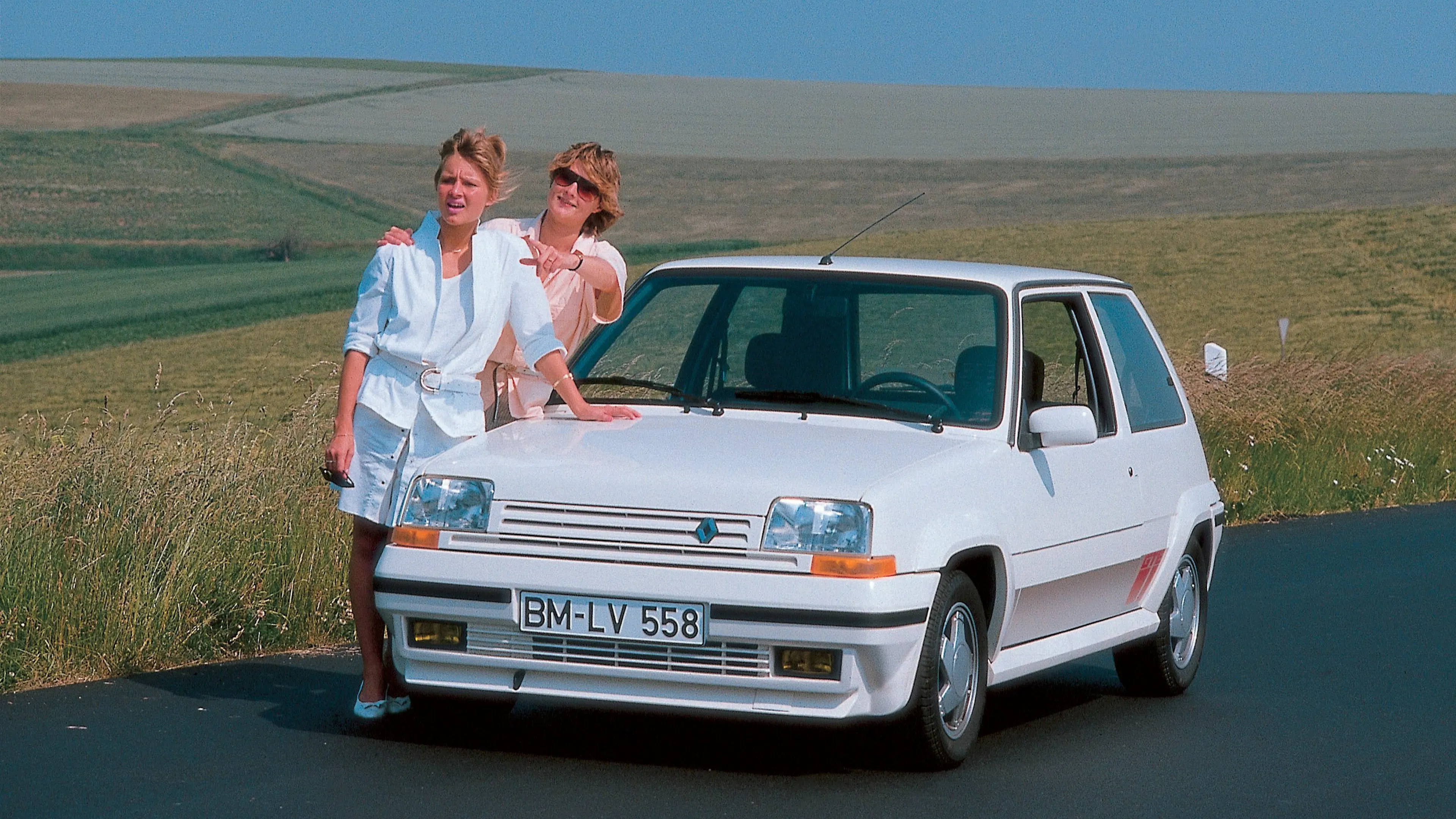 Weißer Renault 5 GT Turbo Phase 2 steht frontal auf einer Straße, zwei Personen lehnen an der Motorhaube.