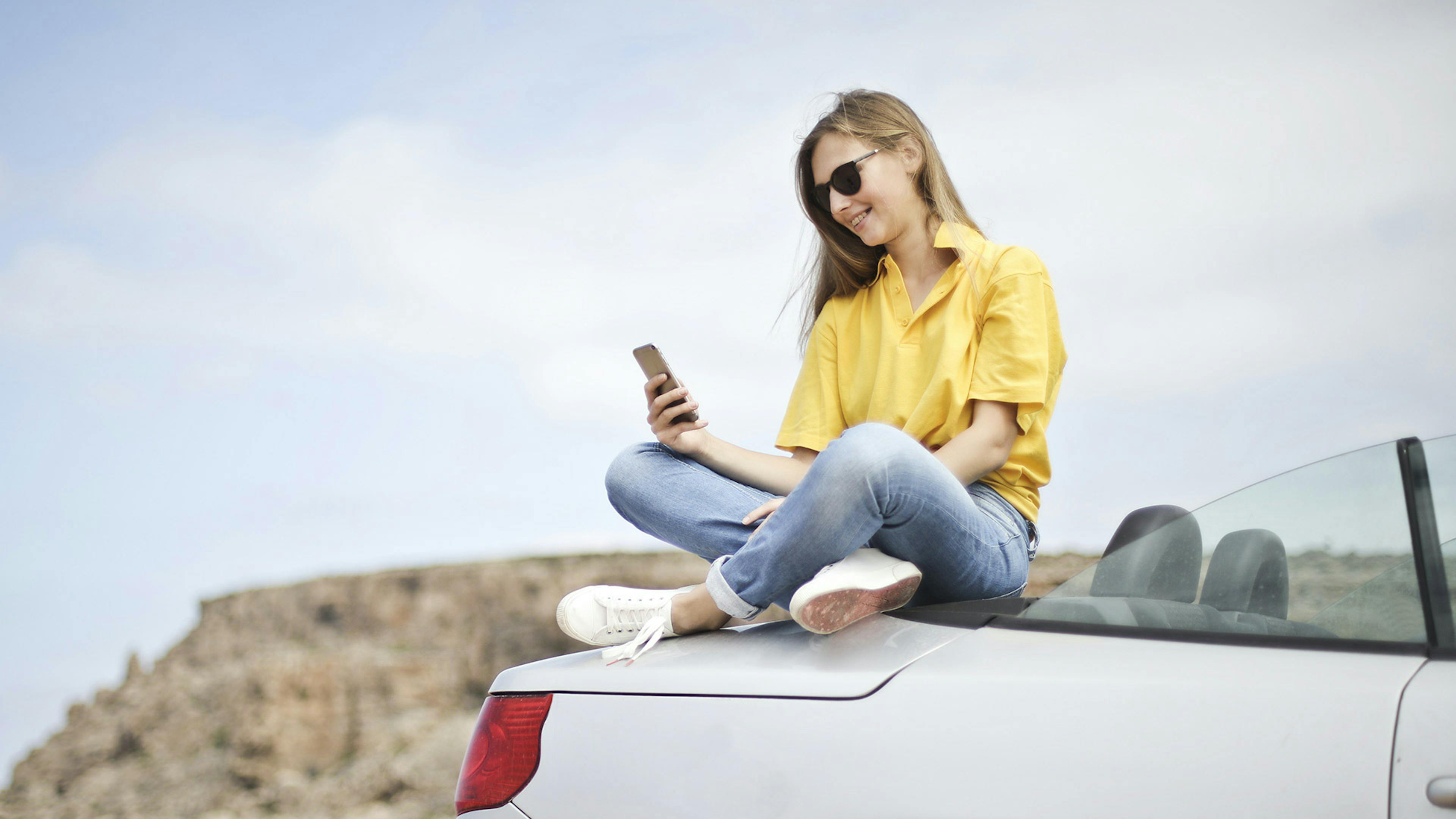 Eine junge Frau sitzt auf dem Heck eines weißen Cabrios und schaut auf ihr Smartphone.