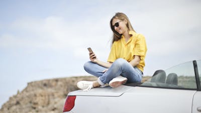 Eine junge Frau sitzt auf dem Heck eines weißen Cabrios und schaut auf ihr Smartphone.