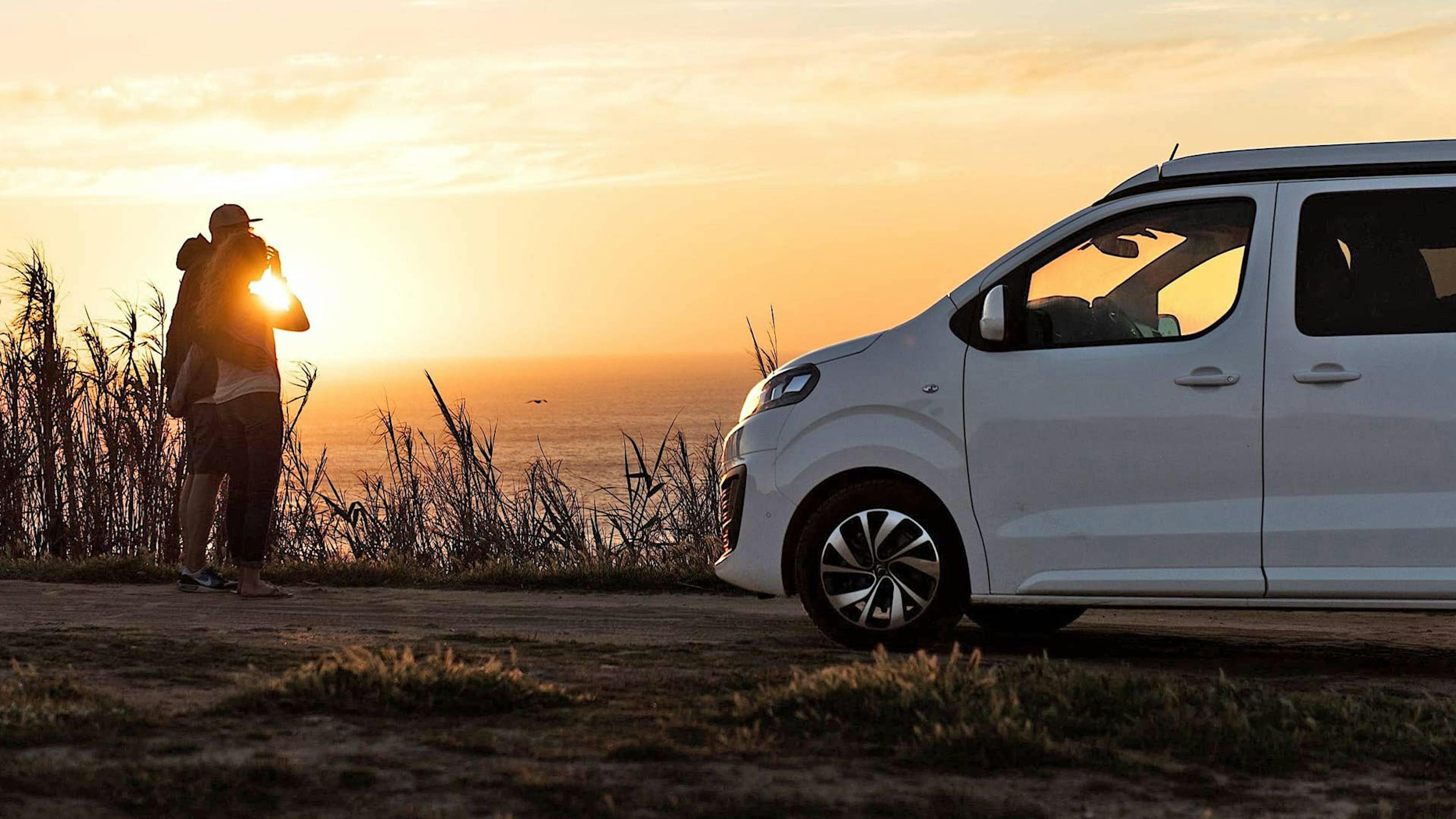 Ein Paar im Urlaub steht am Ufer eines Gewässers neben einem weißen günstigen Wohnmobil und betrachtet einen Sonnenuntergang.